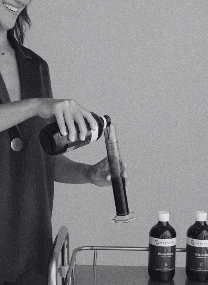 a women pouring herbal liquid from a glass bottle into a graduated cylinder.
