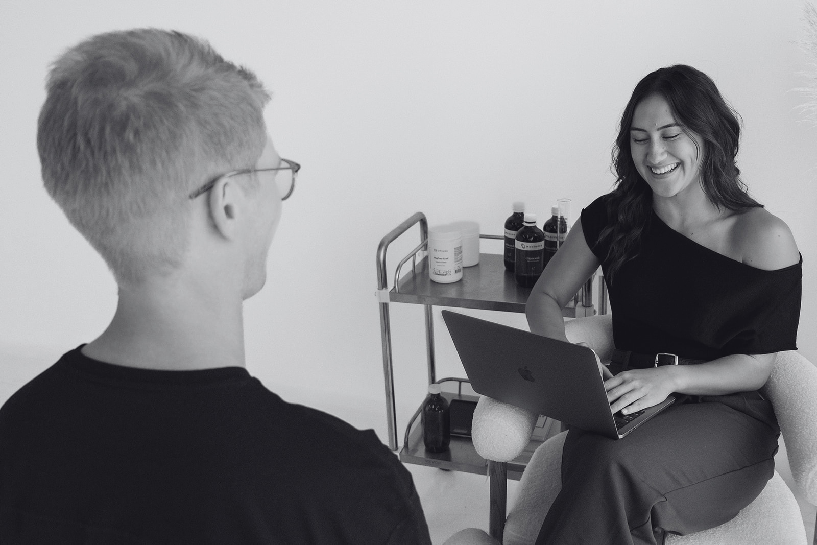 A woman smiling and sitting with a laptop on her lap, talking to a man who is listening. They are in a room with a medical cart containing bottles and containers in the background.