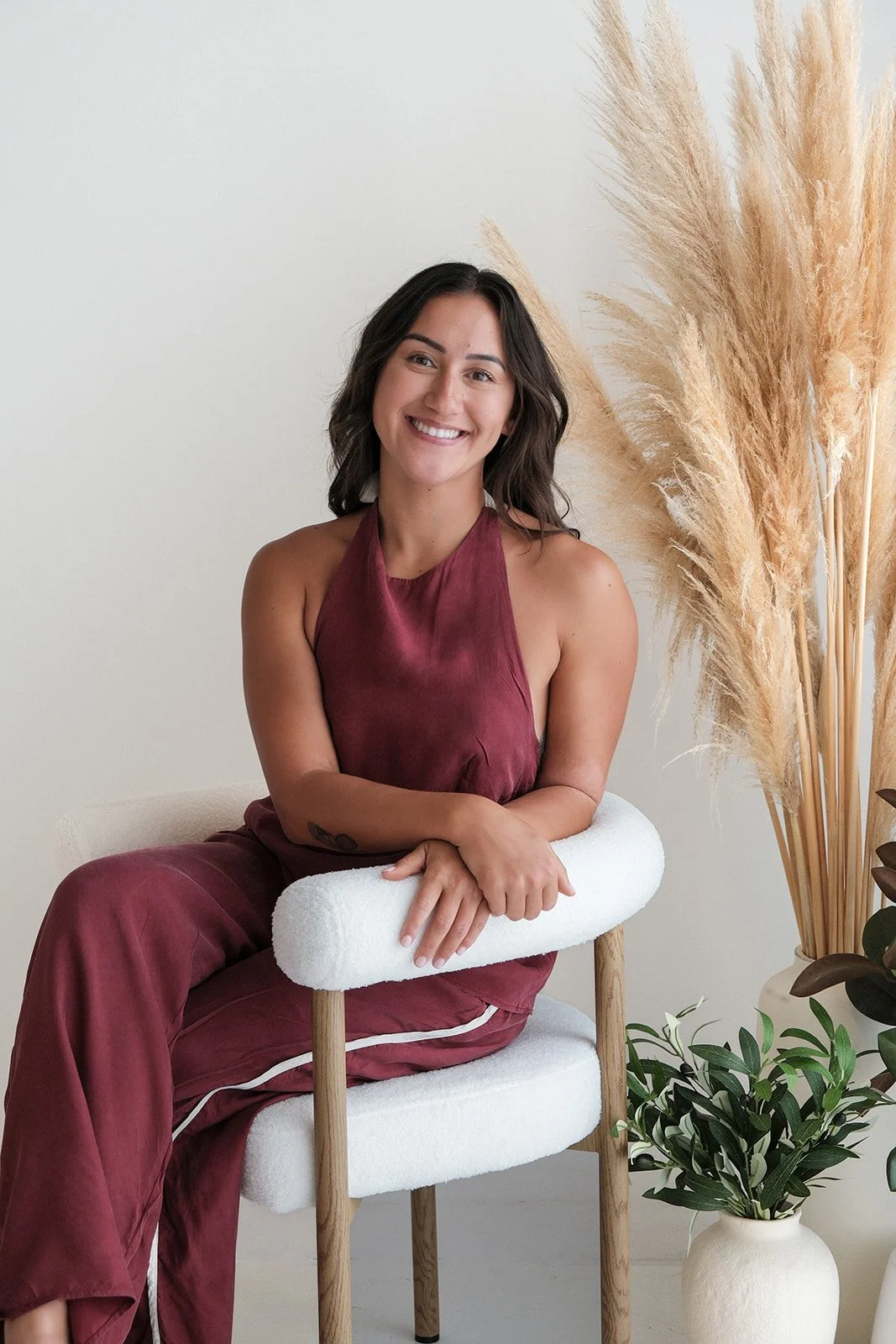 A woman with dark wavy hair smiling, sitting on a modern chair with white cushioning, wearing a sleeveless burgundy top and matching pants, in a minimalistic room with large pampas grass and green houseplants.