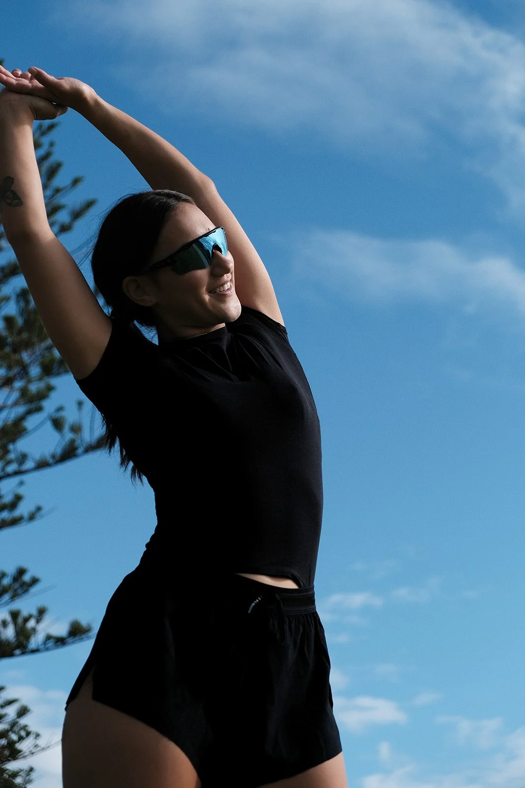 A woman stretching outdoors against a blue sky, wearing sunglasses, a black shirt, and black shorts, smiling.