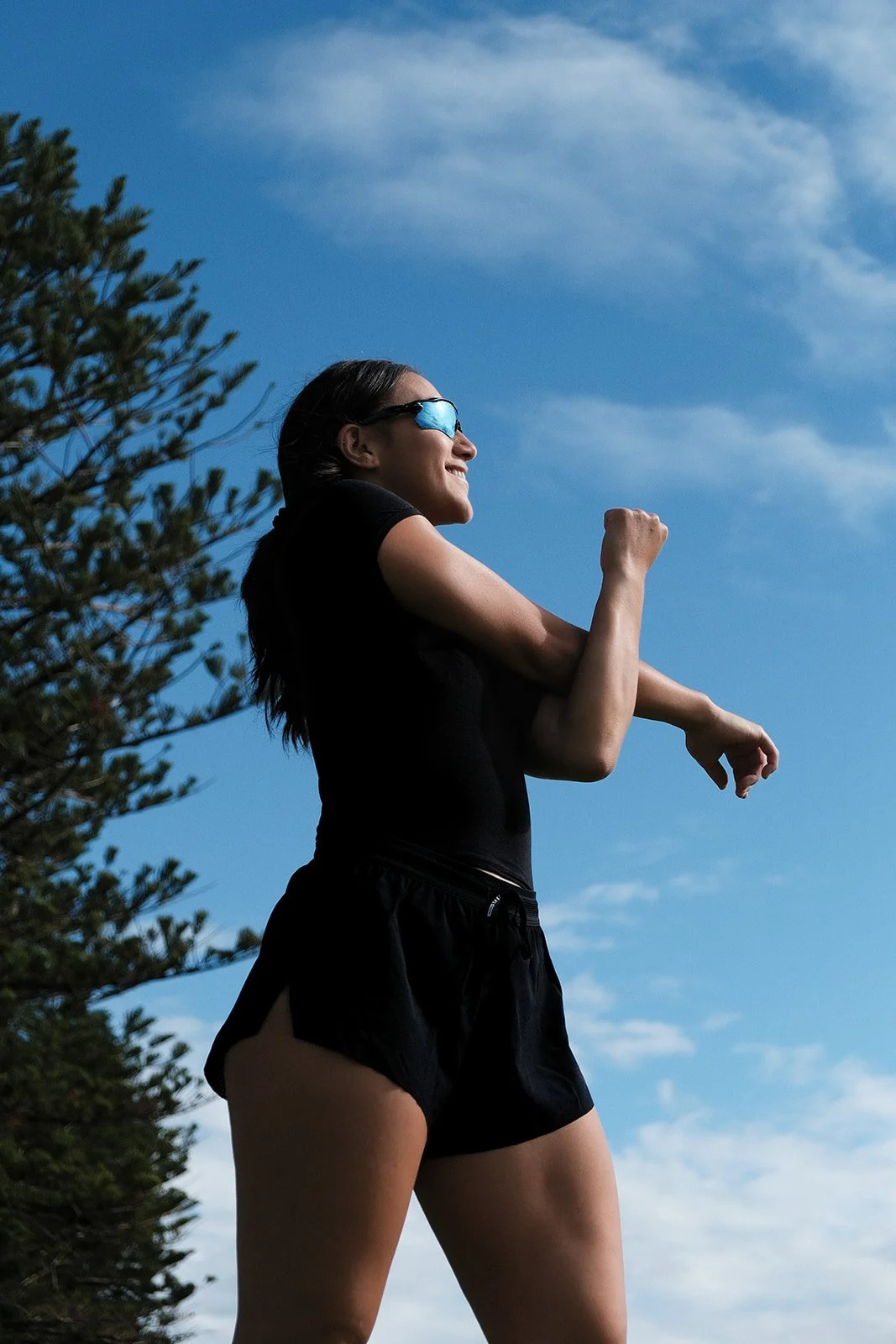 Young woman wearing sunglasses and athletic clothing, smiling and stretching outdoors under a blue sky with some clouds, next to a green tree.