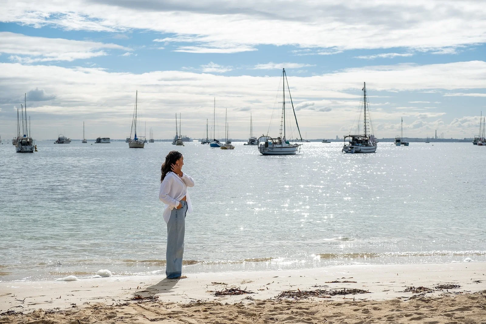 A woman standing on a sandy beach near the water, with her hand behind her head, looking towards sailboats anchored on the calm sea under a partly cloudy sky.