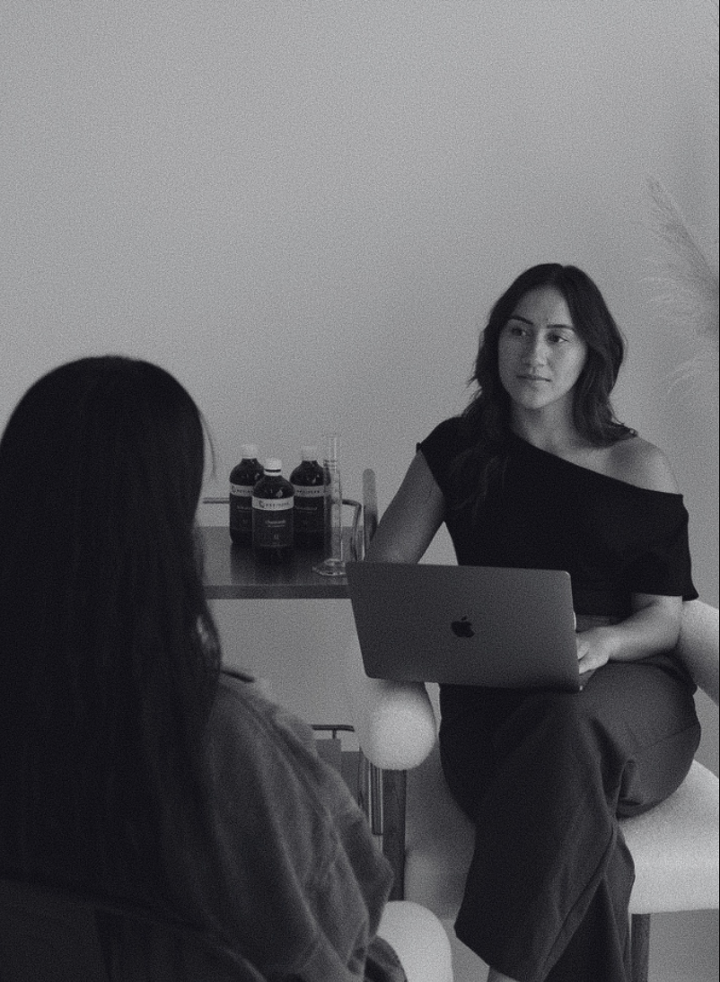 Two women are sitting in a room, engaged in a conversation. One woman is facing forward with a laptop on her lap, while the other woman has her back to the camera. On the table behind them are bottles and a glass, with a plant to the side.