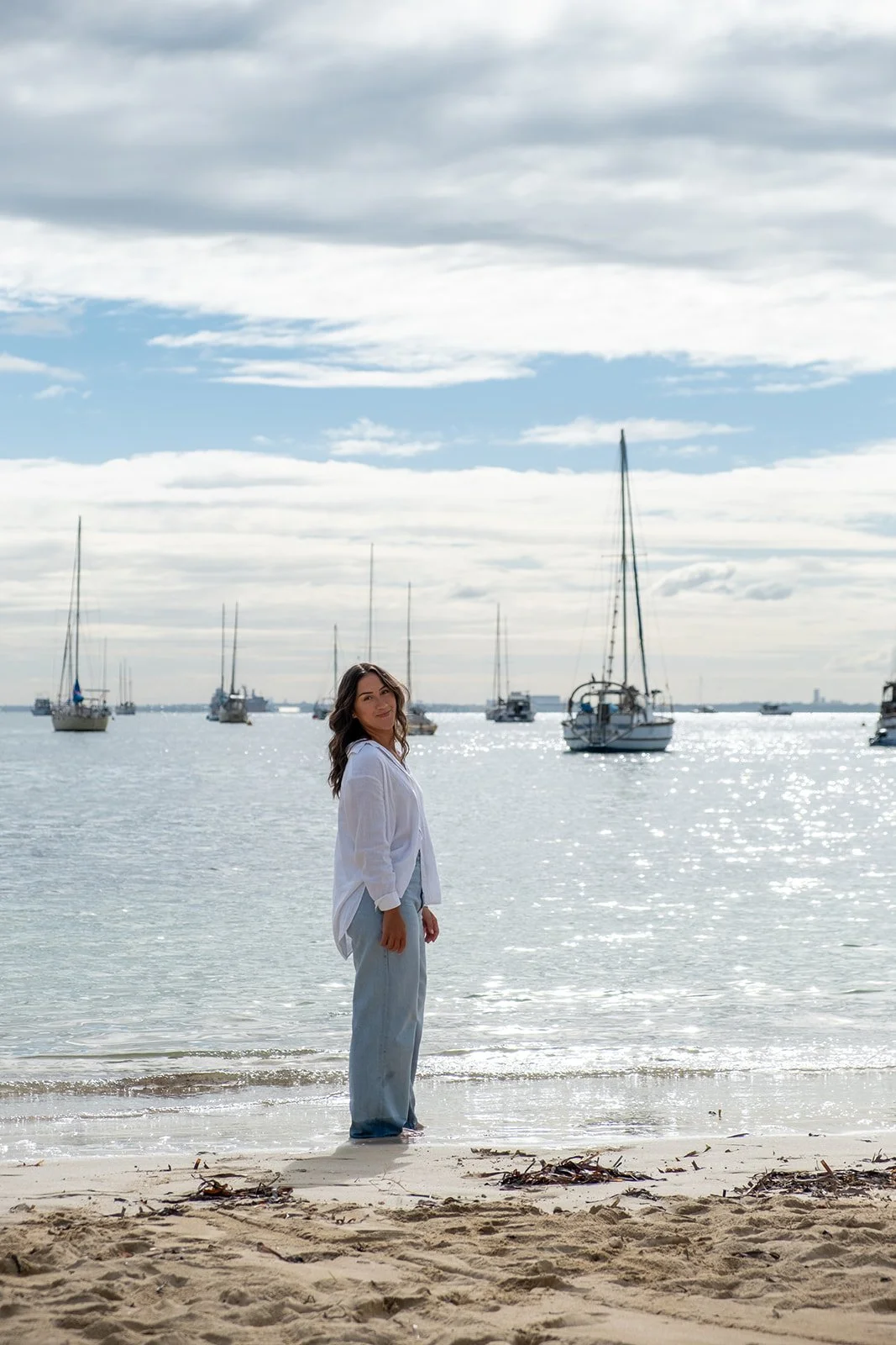 A woman in a white shirt and light blue jeans standing on a sandy beach near the water with sailboats and a cloudy sky in the background.