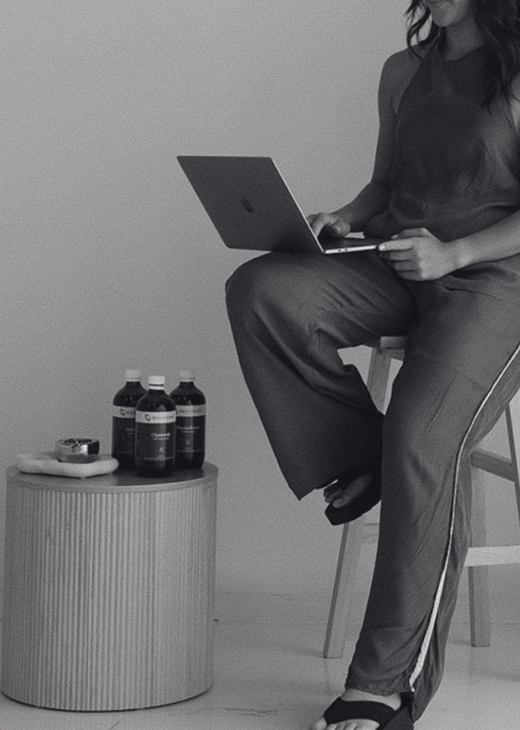 A person sitting on a chair holding a laptop, with three bottles and some items on a small round table nearby.