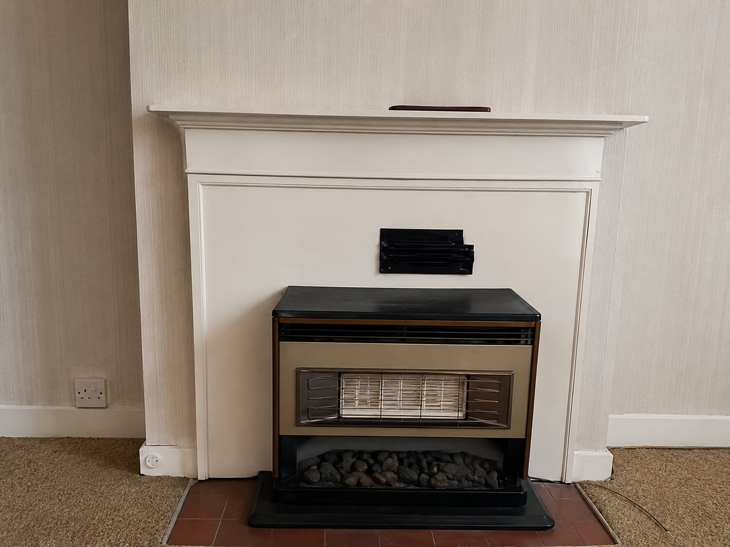 A vintage gas fireplace heater with a black top and brass accents, set into a white wall with a simple white mantel above. There are small rocks in the bottom compartment, and the wall behind is light-colored with textured wallpaper. An electrical outlet is visible to the left of the fireplace, and a power cord runs along the floor.
