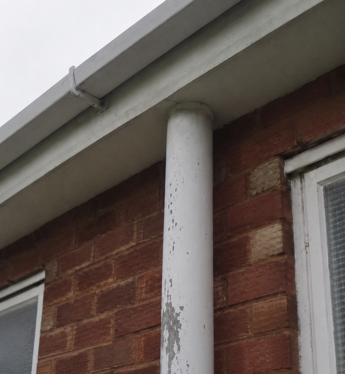 Close-up of the corner of a brick building with a window, showing a white drainpipe and a grey gutter on the roof.