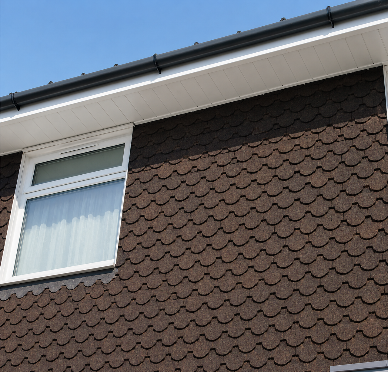 Close-up of the side of a house showing a brown shingle exterior wall with a white-framed window and a dark-colored rain gutter along the roof edge, with a clear blue sky in the background.