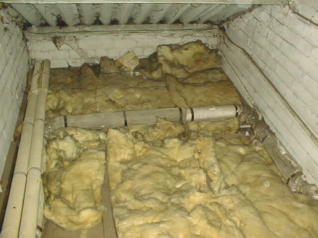 Insulation and plumbing pipes in an attic or crawl space area with exposed brick walls.
