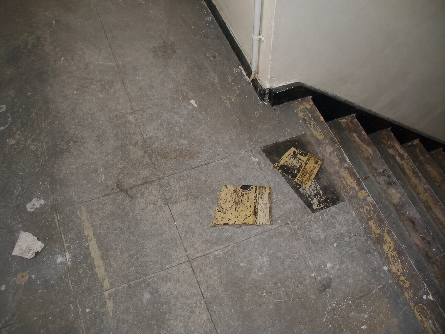Dirty gray tiled floor with peeling paper and debris near a staircase and corner wall.