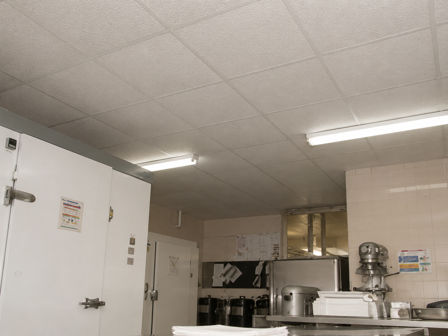 Commercial kitchen with white tiles wall, stainless steel appliances, and overhead fluorescent lights.