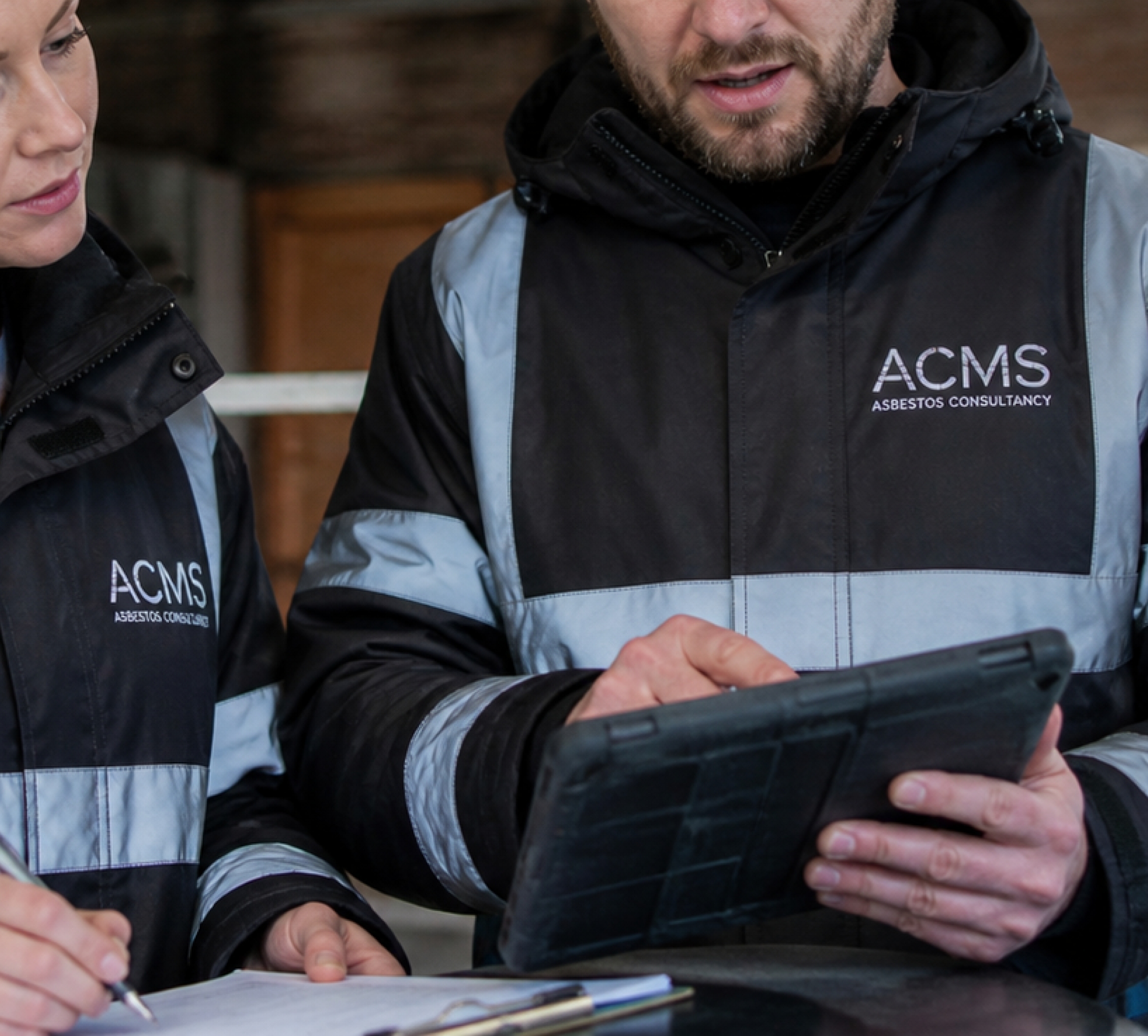 Close-up of two people, one man and one woman, wearing jackets with 'ACMS Asbestos Consultancy' logos, looking at a tablet device together.