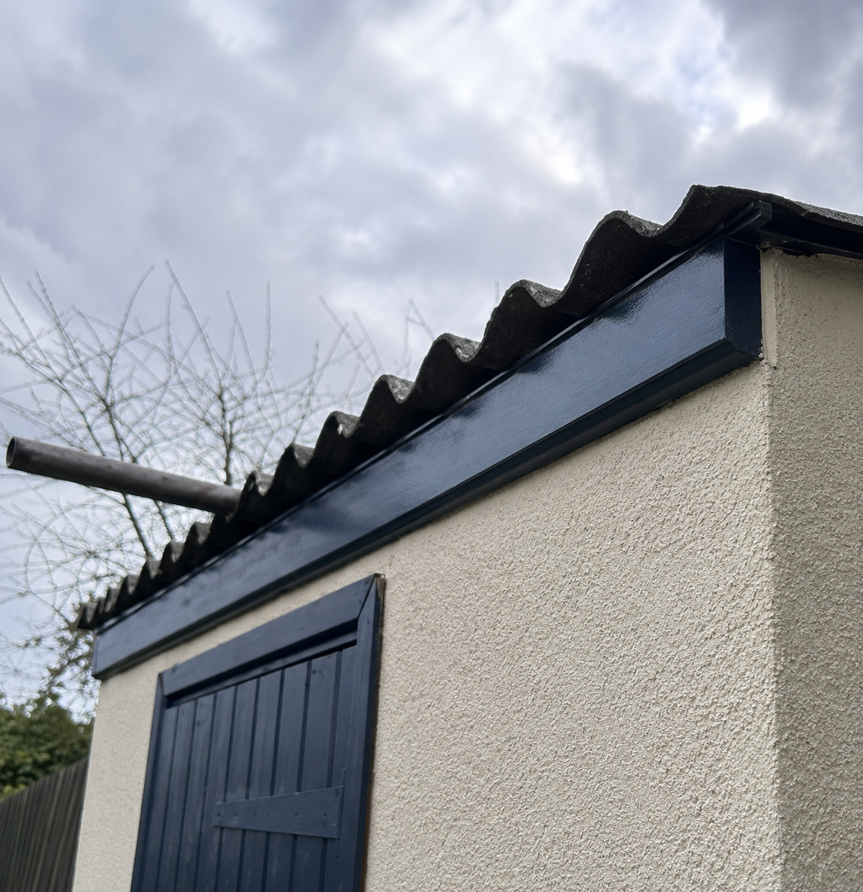 Close-up of a building corner with a beige stucco wall, dark wooden shutters, and a black-painted gutter under a cloudy sky.