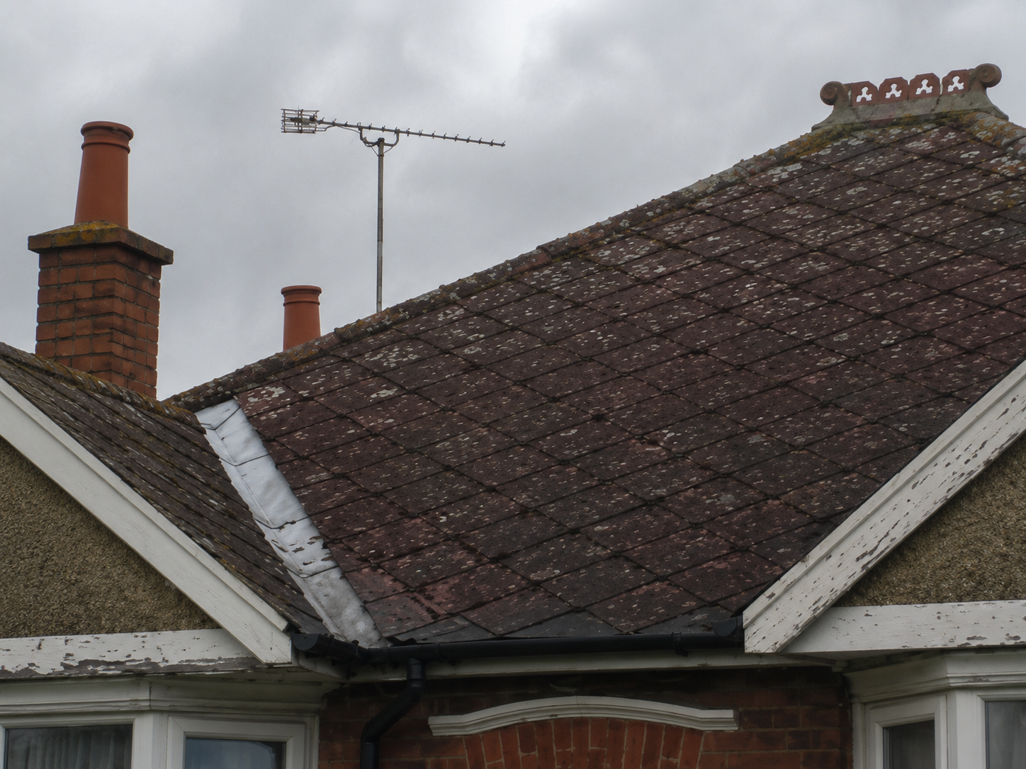 The roof of a residential house with three chimneys, an antenna, and weathered shingles against an overcast sky.
