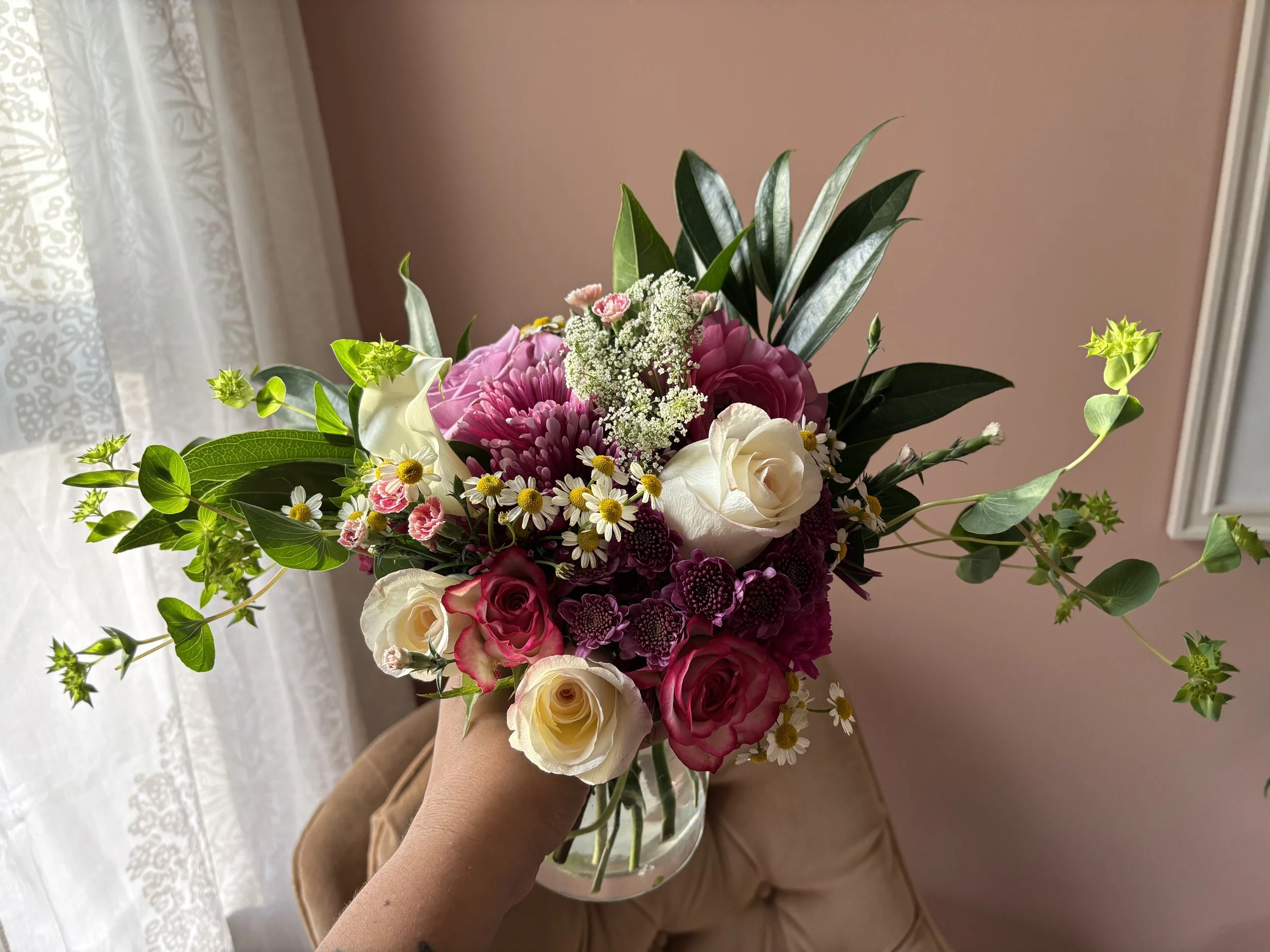 Close-up of a colorful bouquet of flowers held by a hand, with a pink wall and white curtain in the background.