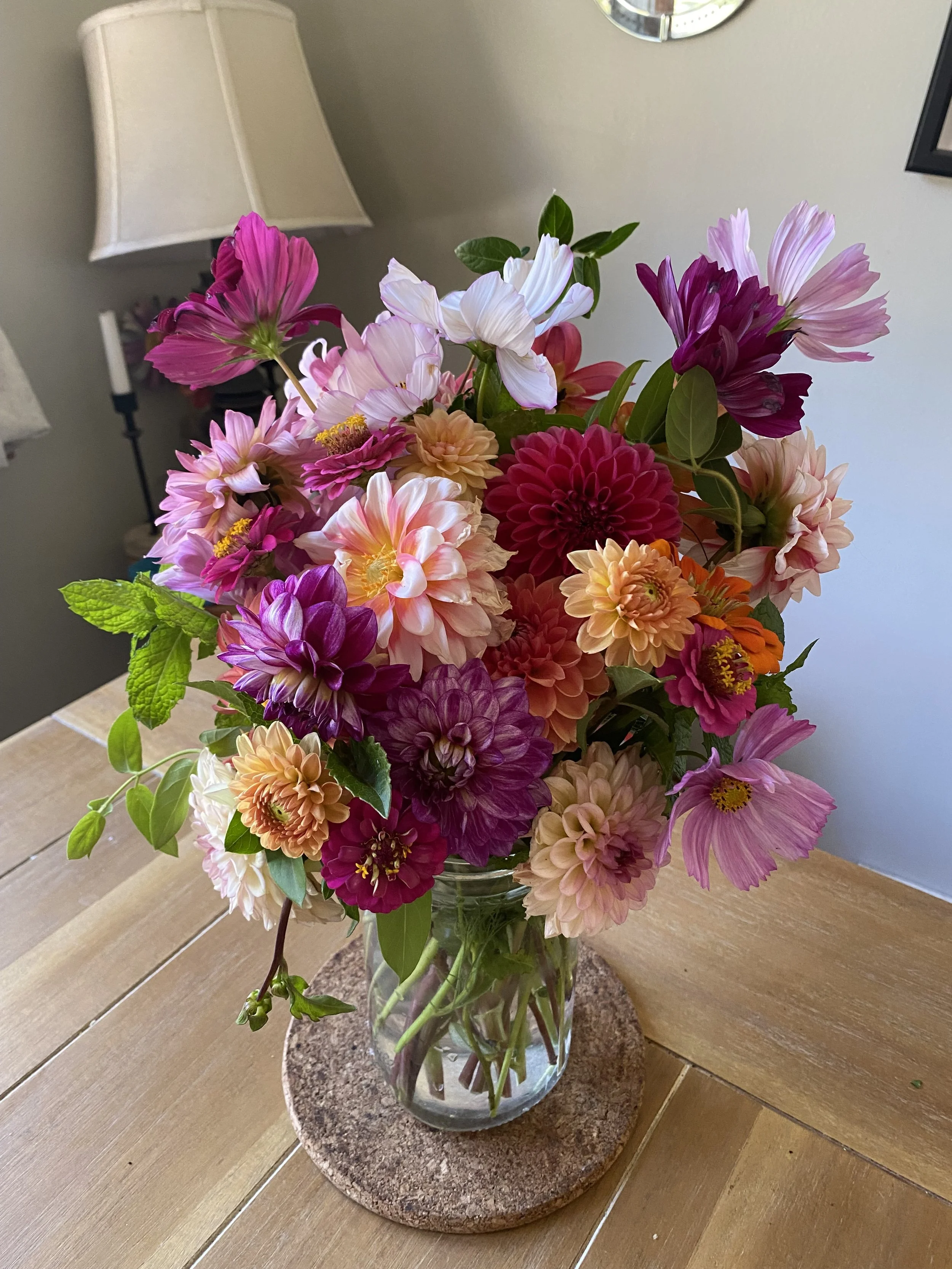 A bouquet of colorful flowers in a clear glass vase on a wooden table with a granite coaster underneath, a lamp, and a mirror in the background.