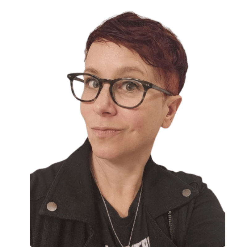 A person with short red hair, glasses, and a playful expression, wearing a black jacket and a necklace.