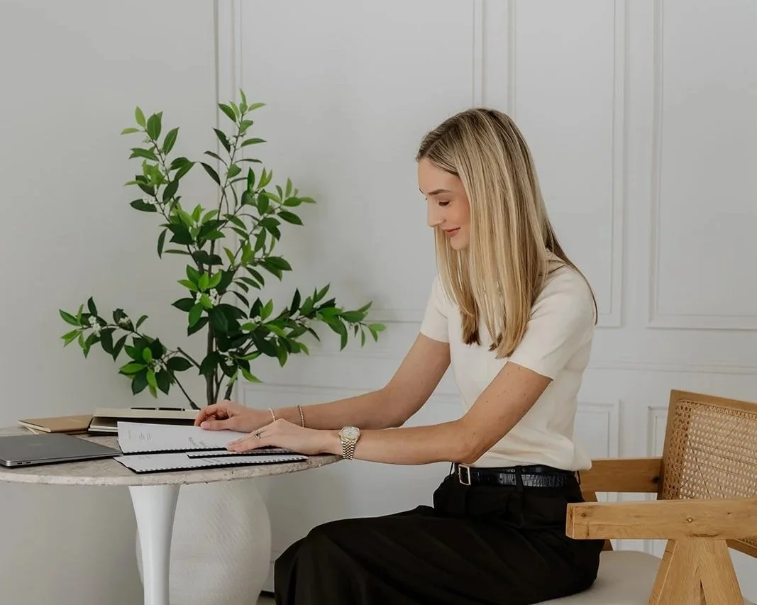 Founder Rebecca Simmons sitting at a round table in a minimalistic room, reading an estate planning document, with a large green plant and a laptop on the table.