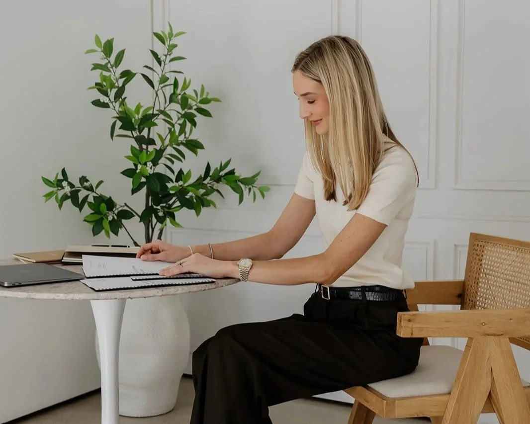 A melbourne lawyer with blonde hair, wearing a white shirt and black pants, sitting at in the Melbourne office, reading a book with a laptop and a plant nearby.