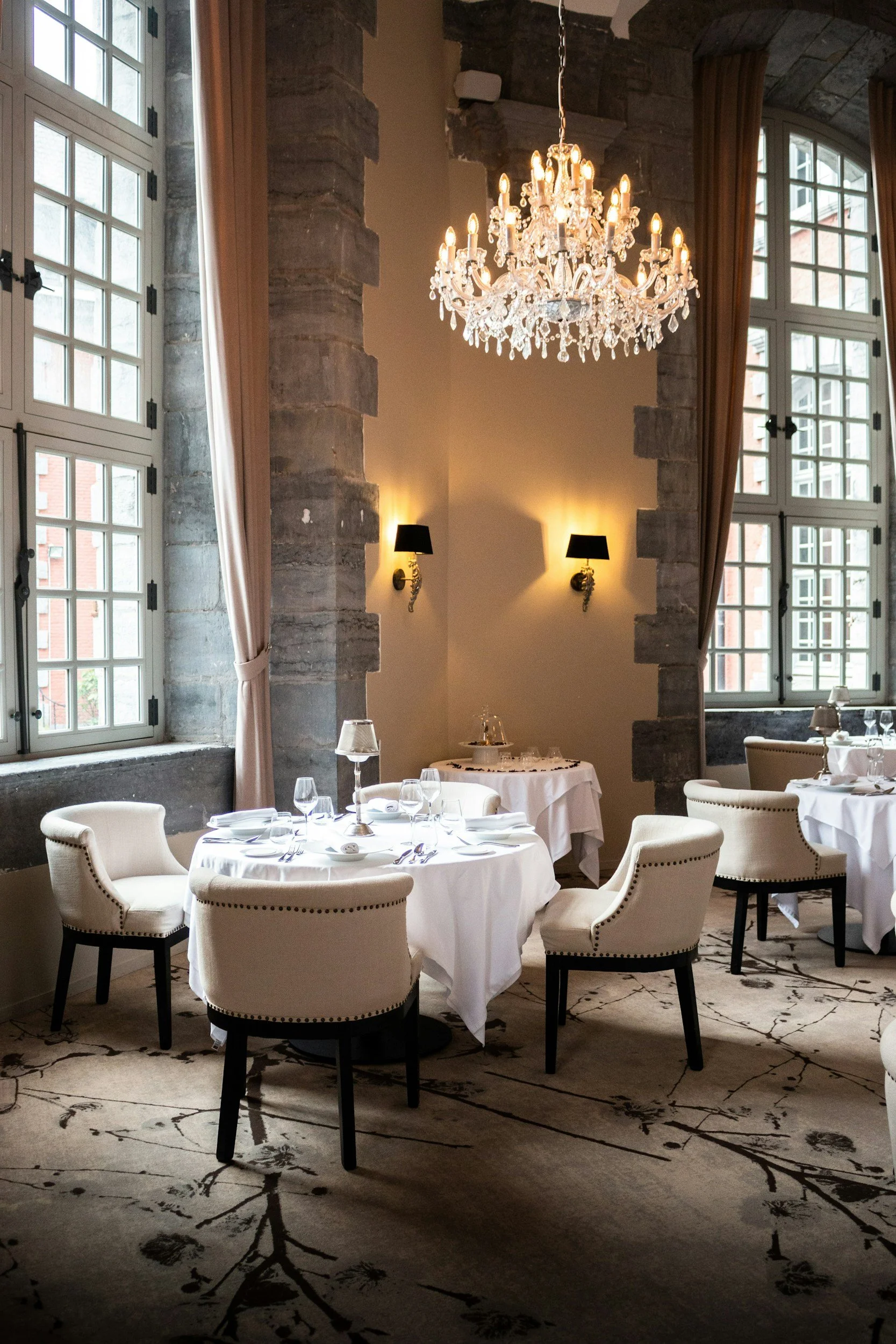Elegant dining room with round tables set with white tablecloths, chairs with cream upholstery, large windows with curtains, marble floor with black branch and flower pattern, stone walls, chandelier, and wall sconces.