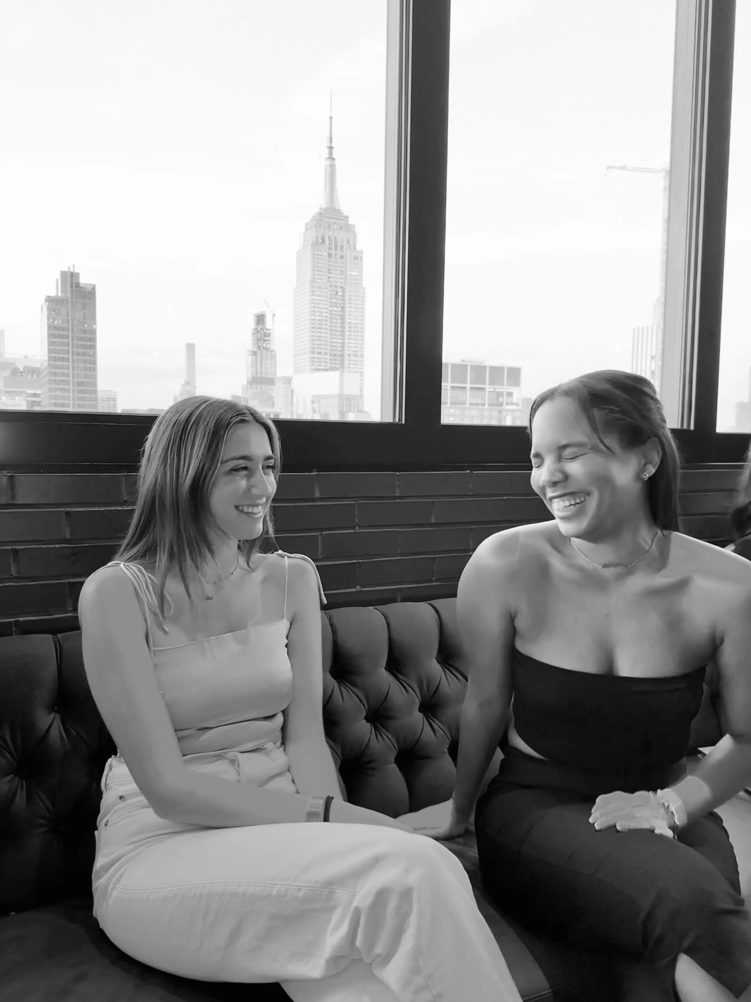 Two women sitting on a tufted leather sofa near large windows with a city skyline in the background, smiling and engaging with each other.