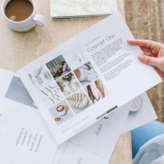 Person holding a brochure or magazine labeled 'concept one' with interior design images, a cup of coffee on a table, and other papers or magazines.