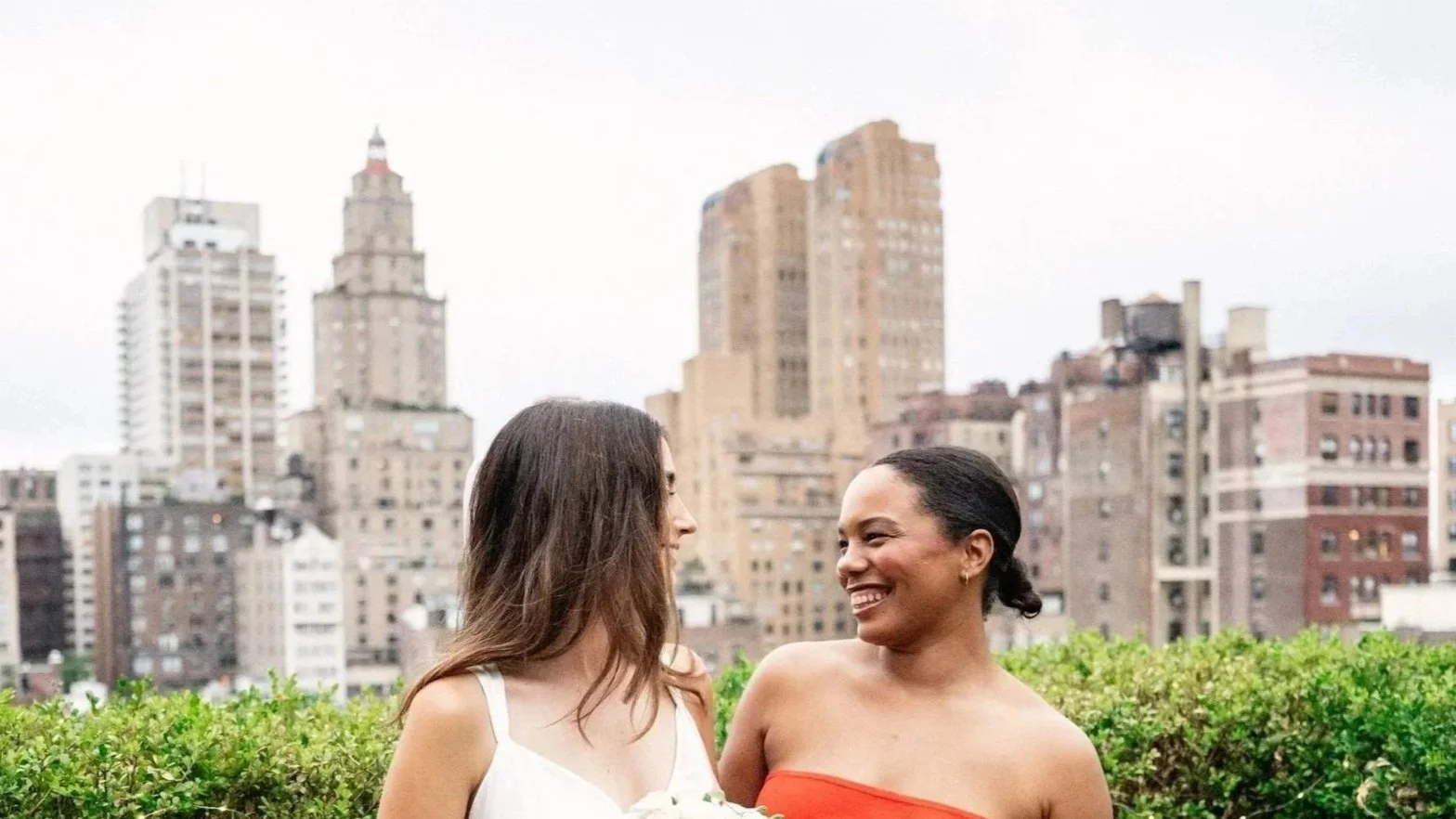 Two women smiling at each other outdoors on a rooftop with a city skyline in the background.