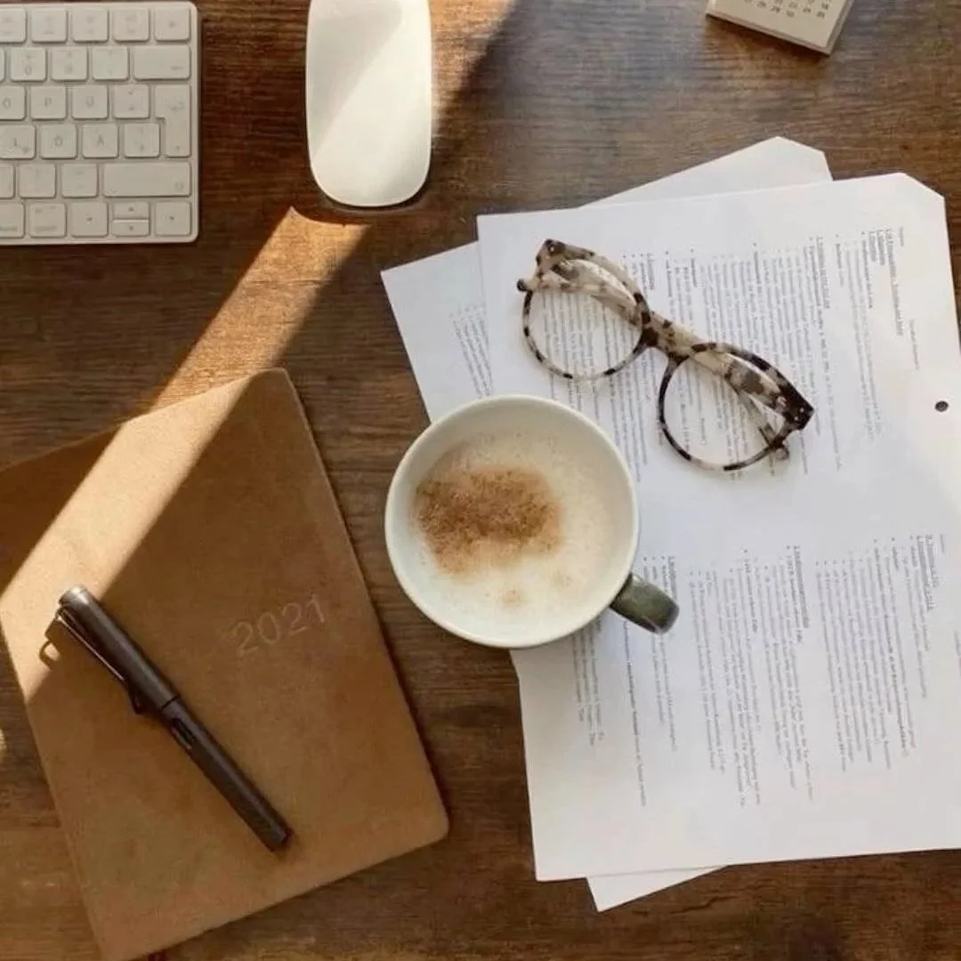 A wooden desk with a white keyboard, a white computer mouse, a brown 2021 planner, a black pen, a cup of coffee with foam and cinnamon, a pair of glasses with a tortoiseshell frame, some papers, and a calculator.