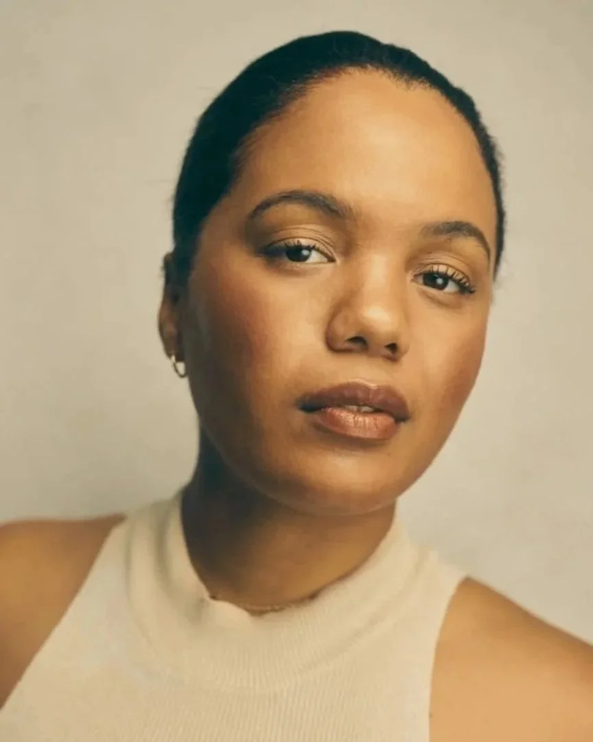 Close-up portrait of a woman with short, dark hair, wearing a sleeveless beige turtleneck top, with a neutral expression and minimal makeup.