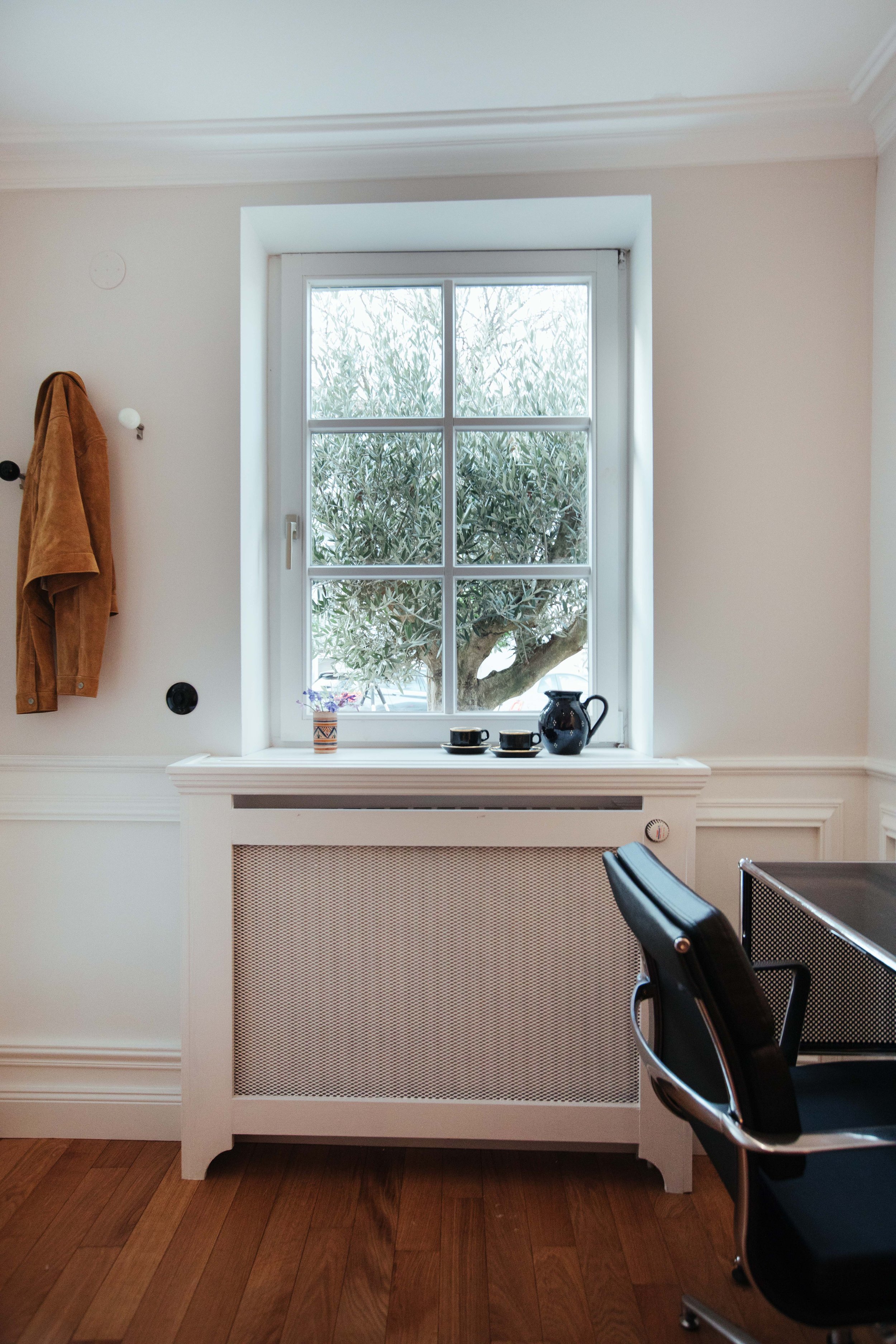 Ein Fenster mit Blick auf einen Baum mit grünen Blättern, eine Vase mit Blumen, eine Kanne und zwei Tassen auf der Fensterbank, ein Stuhl und ein Radiator darunter, weiße Wände.