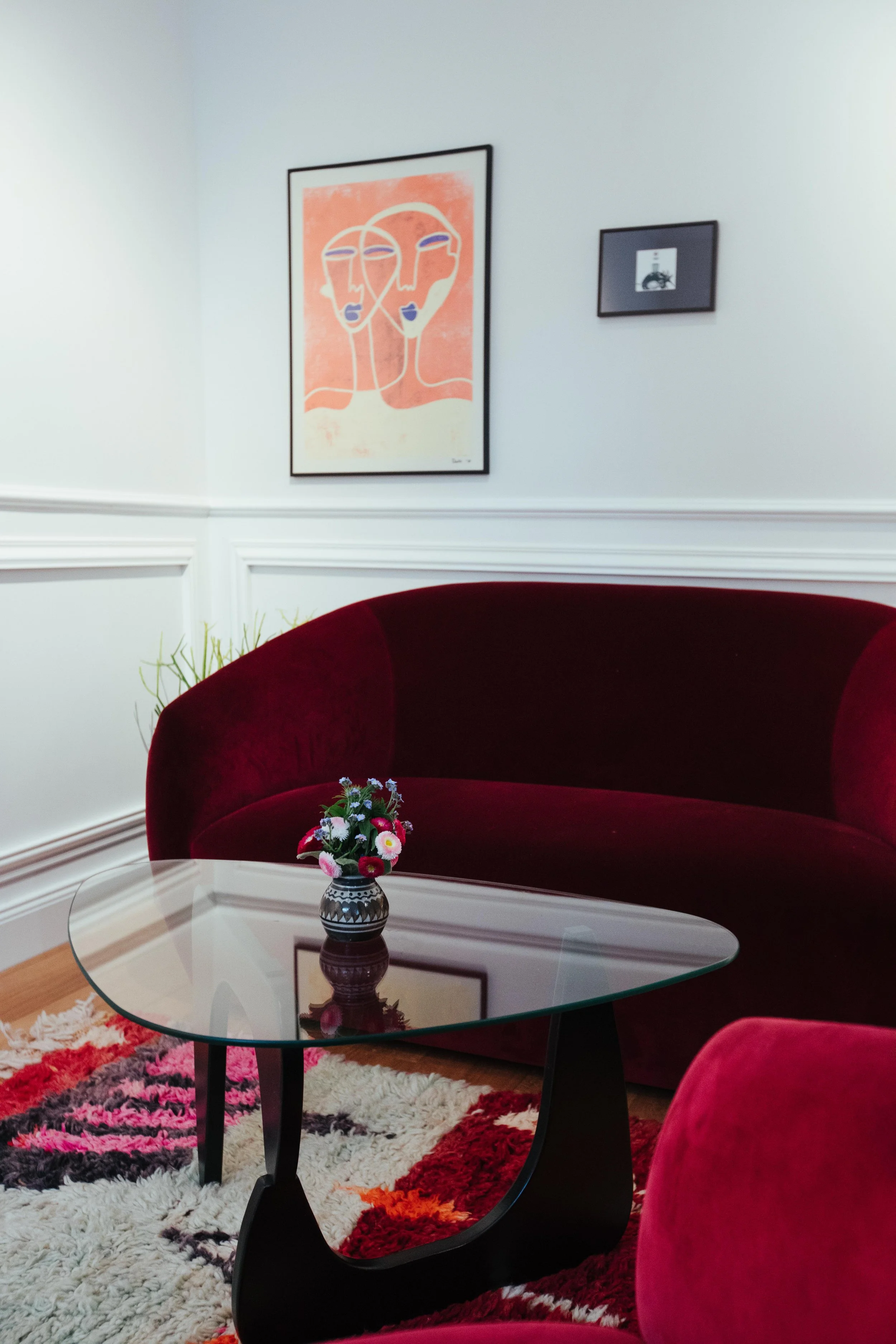 Ein rotes Sofa, ein Glastisch mit Vase und Blumen, weiße Wand mit zwei Kunstwerken, ein Teppich mit buntem Muster.