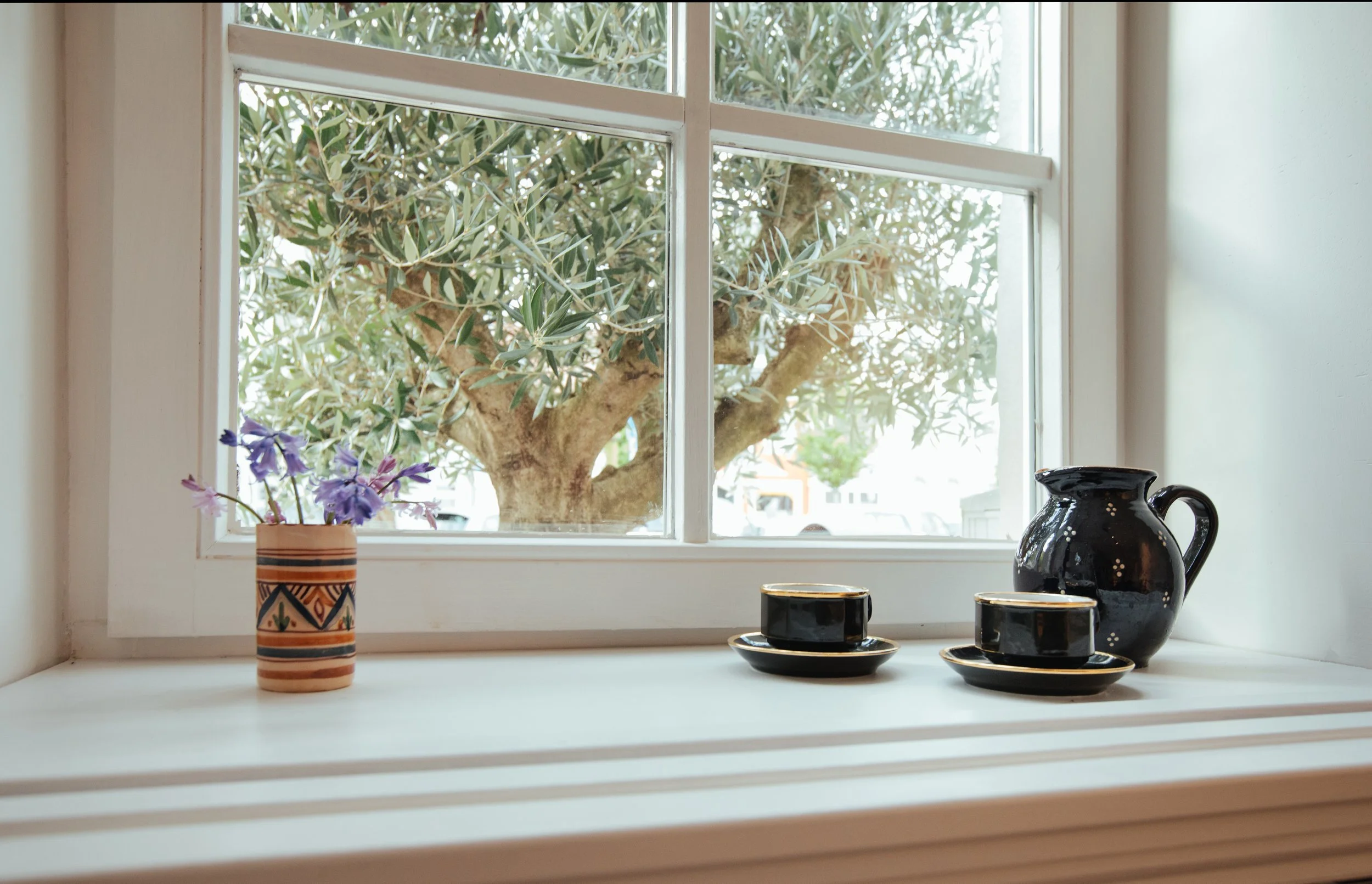 Blick durch eine Fensterbank auf eine schwarze Keramik Karaffe, einen kleinen schwarzen Teller und eine Kaffetasse. Auf der Fensterbank steht eine bunte Keramikvase mit blau-violetter Blume. Hinter dem Fenster sieht man einen Baum mit grünen Blättern.