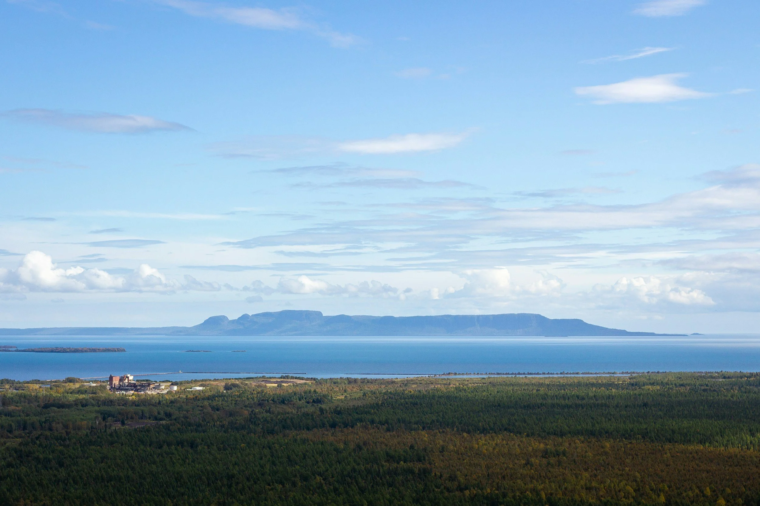 A landscape with a large body of water, distant mountains, and green forests in the foreground under a partly cloudy sky.