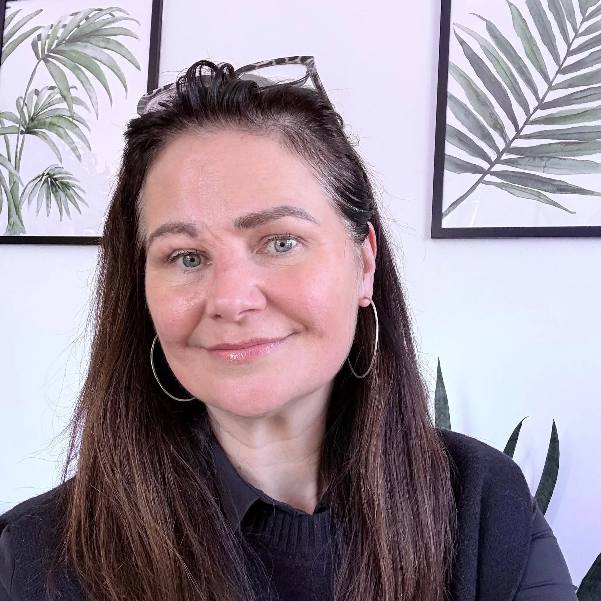 A woman with long, dark hair and light skin is smiling slightly, looking into the camera. She is wearing a black top and earrings. Behind her, there are two framed botanical art prints of green fern leaves on a white wall.