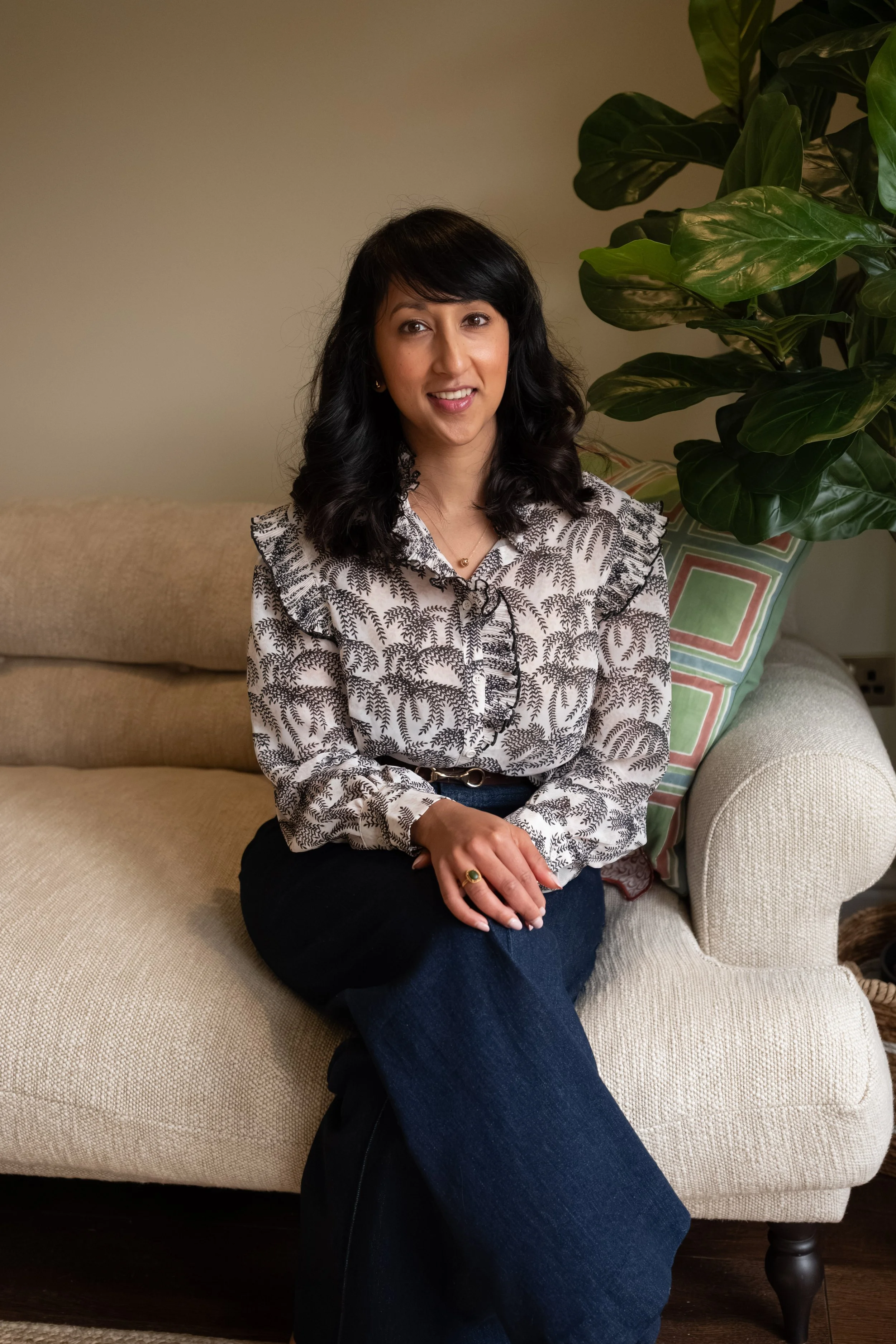 A woman with curly black hair sitting on a beige sofa, wearing a patterned blouse and dark jeans, with a large leafy houseplant behind her and a colorful pillow nearby.