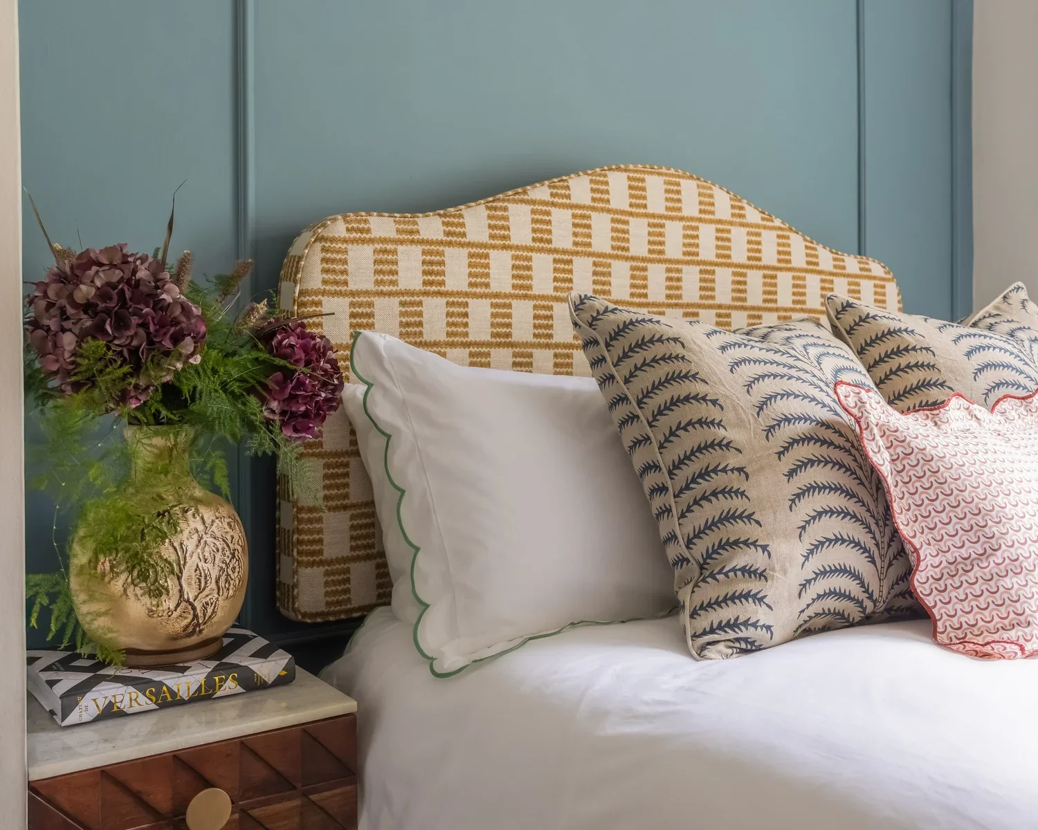 A bedroom scene featuring a bed with a patterned headboard, decorative pillows, a floral arrangement in a gold vase, and a book titled 'Versailles' on a bedside table.