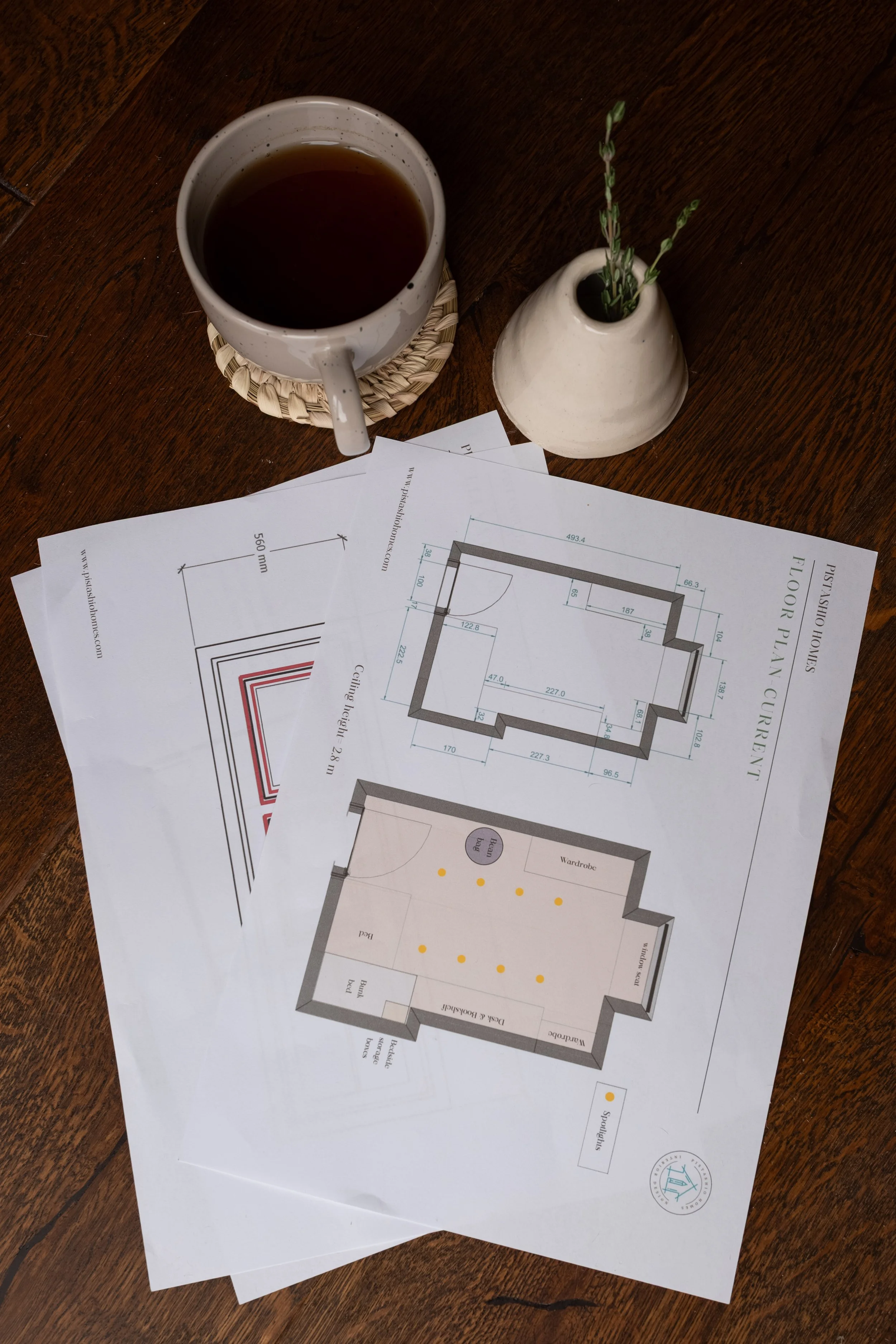 A wooden table with rolled architectural floor plans, a white cup of tea on a woven coaster, and a small white vase with green sprigs.