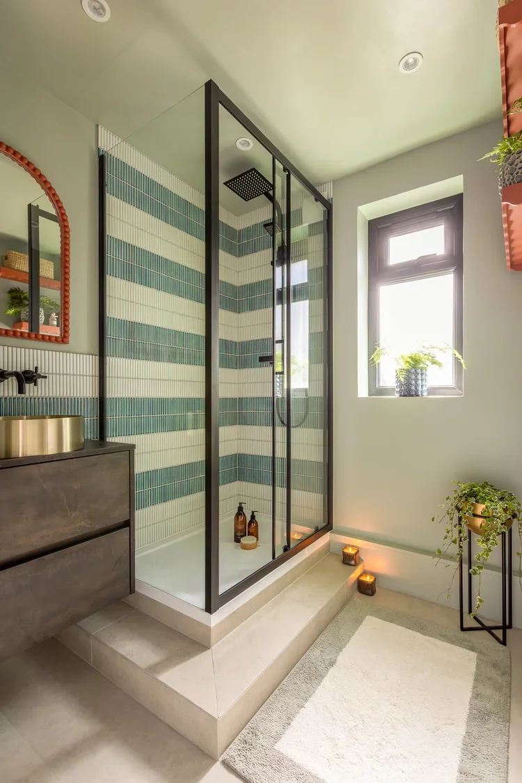 A modern bathroom featuring a glass-enclosed shower with horizontal striped teal and white tiles, a window with a potted plant, a vanity with a mirror, and candles on the floor.