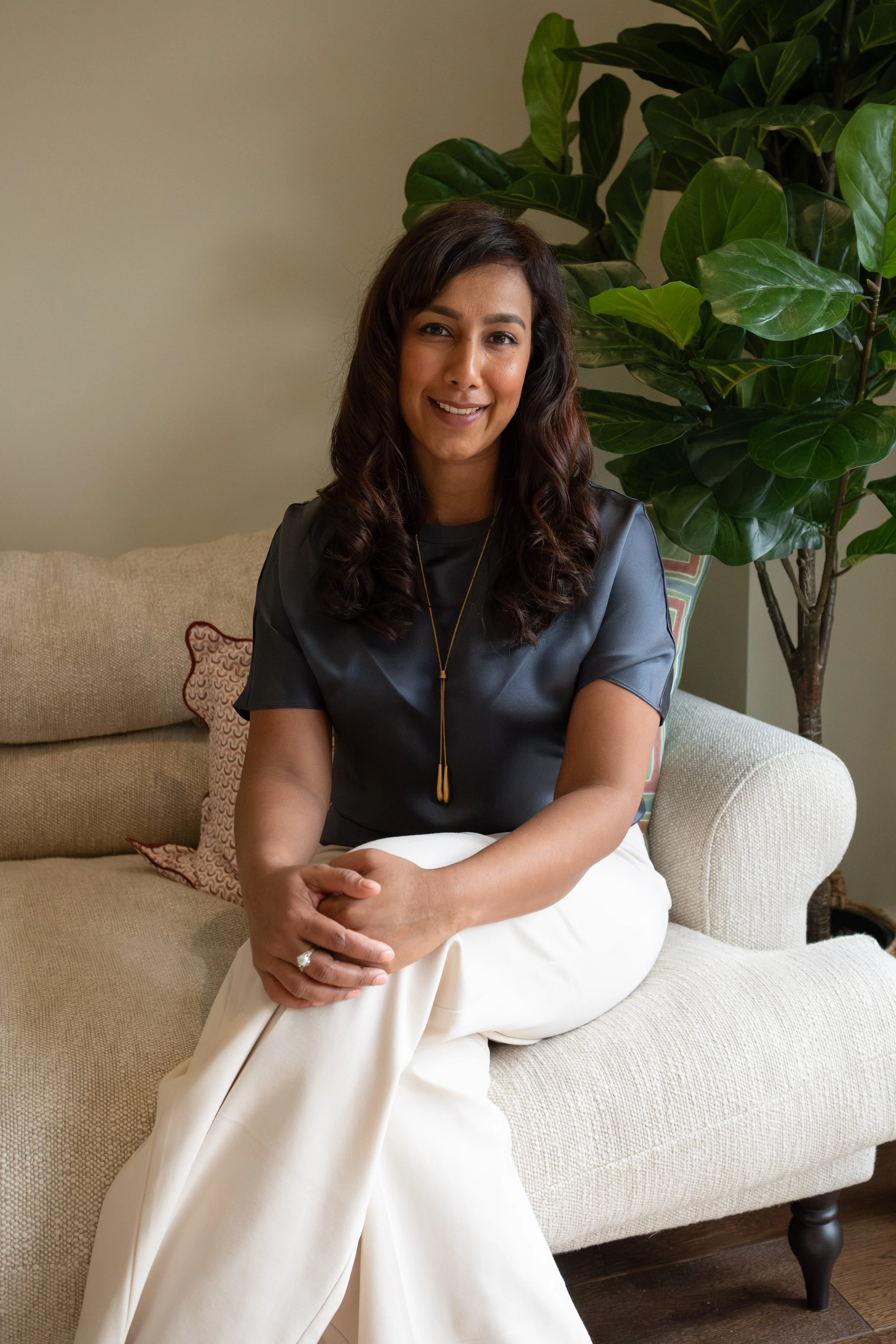 A woman with dark wavy hair and a warm smile, sitting on a beige sofa with decorative pillows, beside a tall green leafy plant in a cozy room.