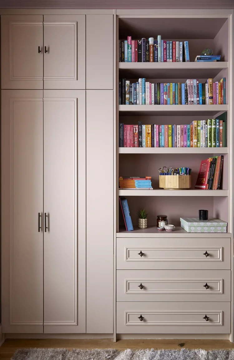 A bookshelf and cabinet unit with pinkish-beige color. The bookshelf contains colorful books, a small potted plant, and various decorative items. The cabinet has closed doors and three drawers with metal knobs.