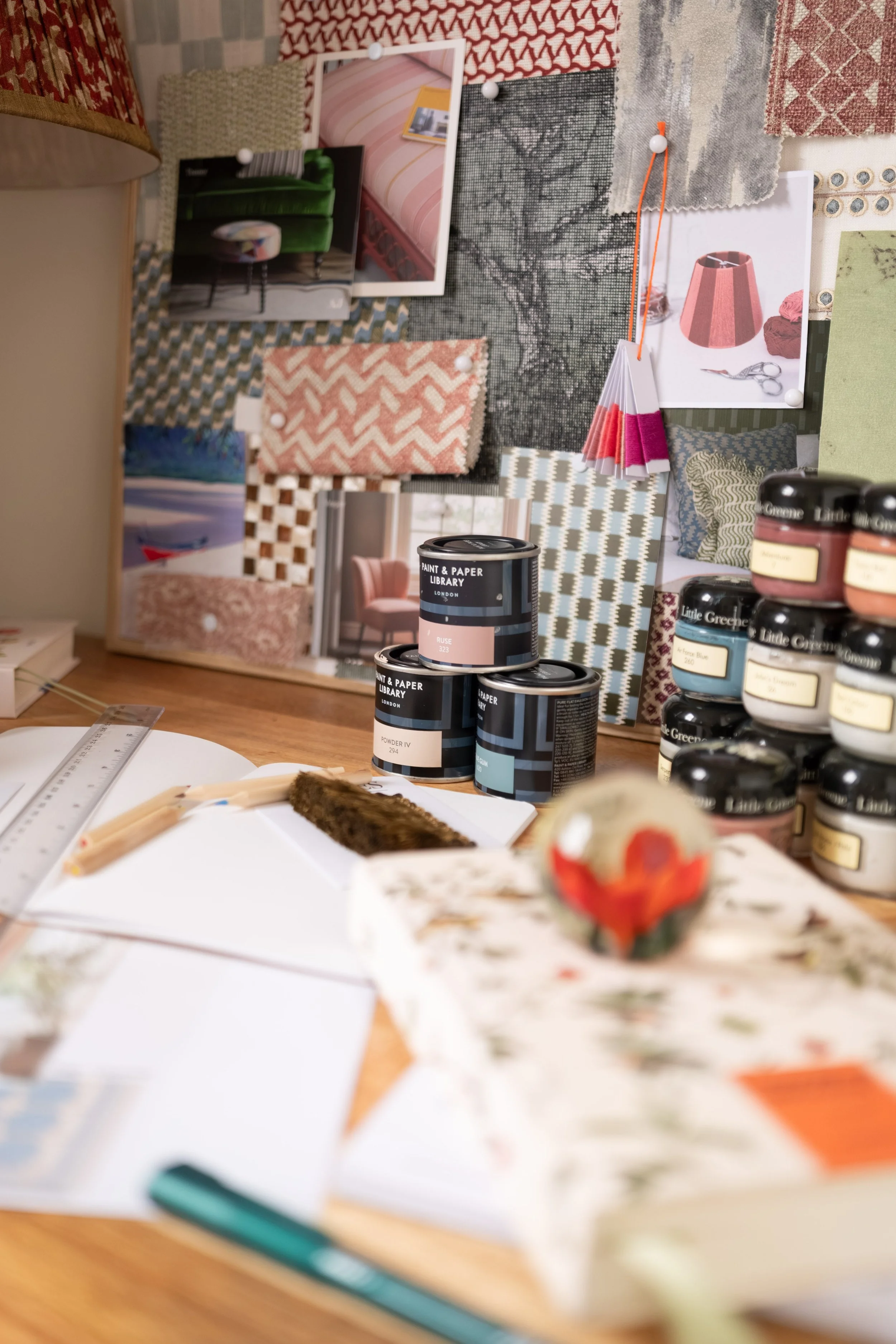 A close-up view of a craft workspace with paint jars stacked on a wooden table, a paint swatch fan deck, and a blurred background of fabric samples and photographs pinned to a board.