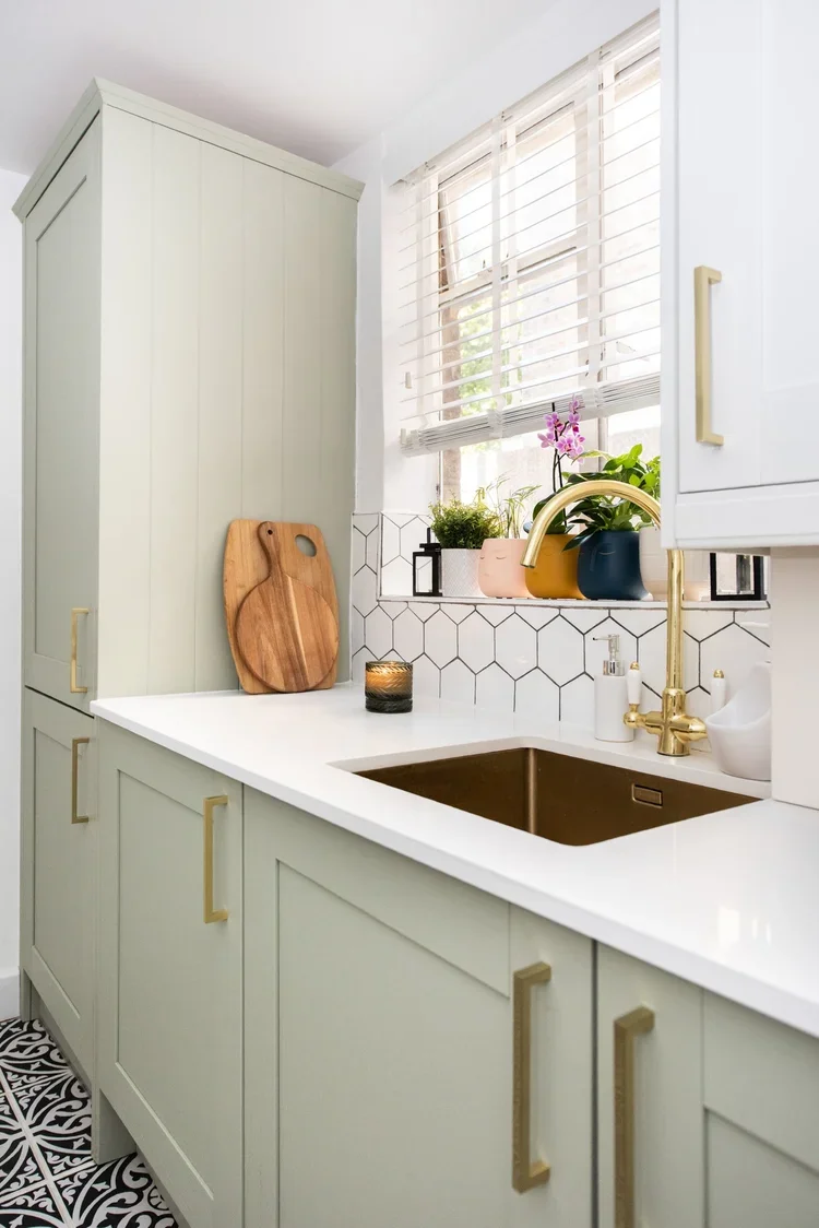 A kitchen with pale green cabinets, a white countertop, a gold faucet, and a dark brown sink. There are potted plants and a purple orchid on the window sill, white window blinds, and a patterned tile floor.