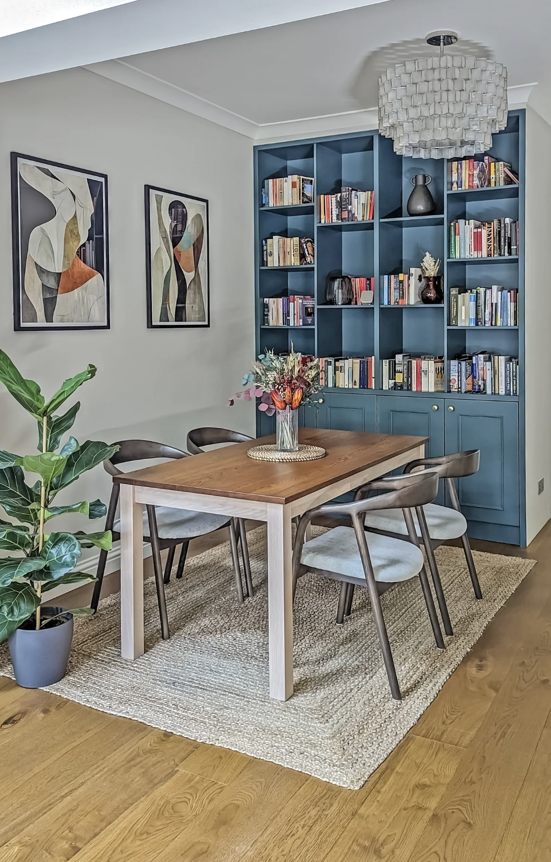 Contemporary dining room with a wooden table, six chairs, a blue built-in bookshelf, a vase of flowers, artwork on the wall, a potted plant, and a modern chandelier