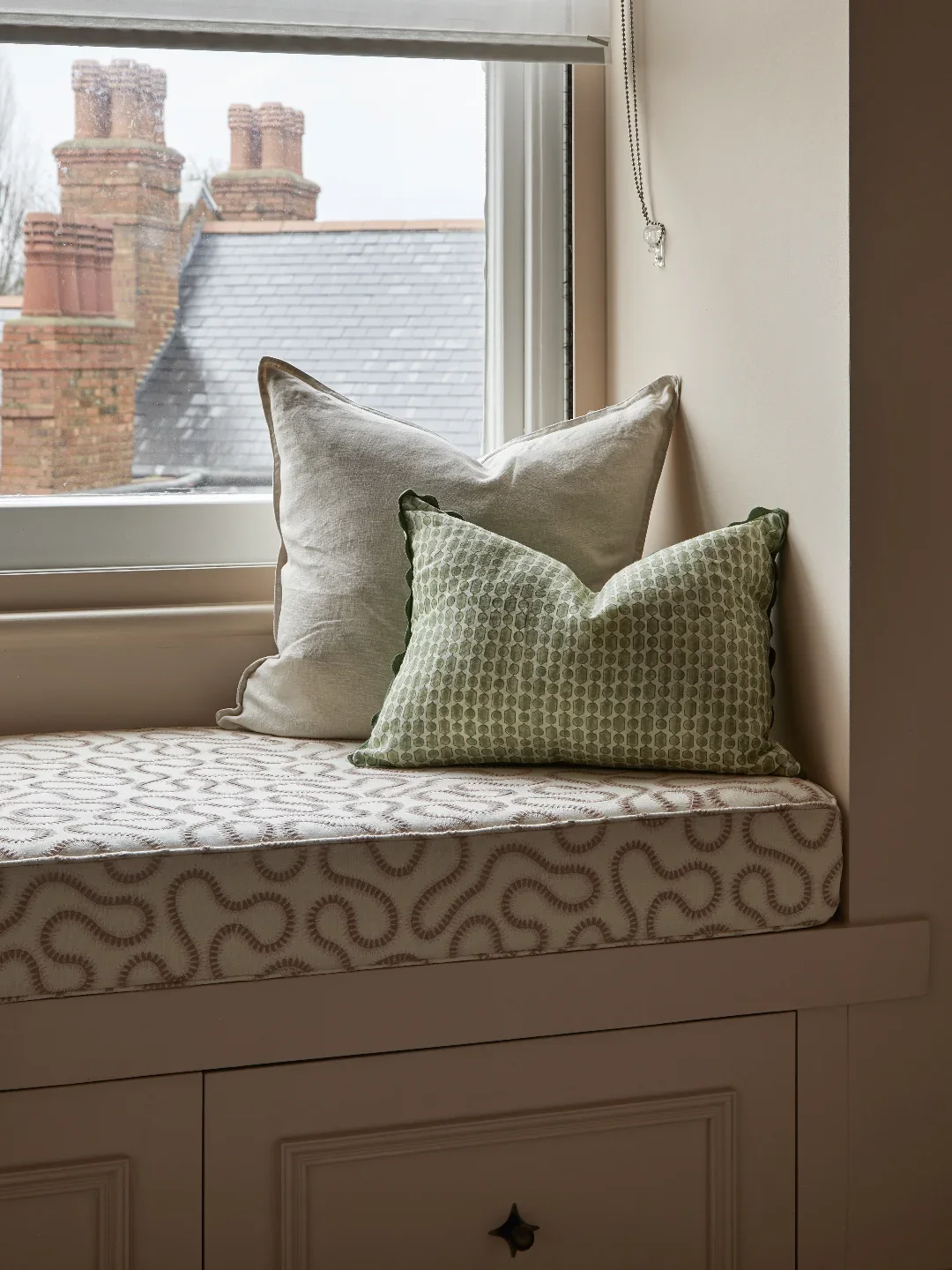 A window seat with two decorative pillows, one beige and one patterned green, near a window with a view of brick building chimneys and rooftops.