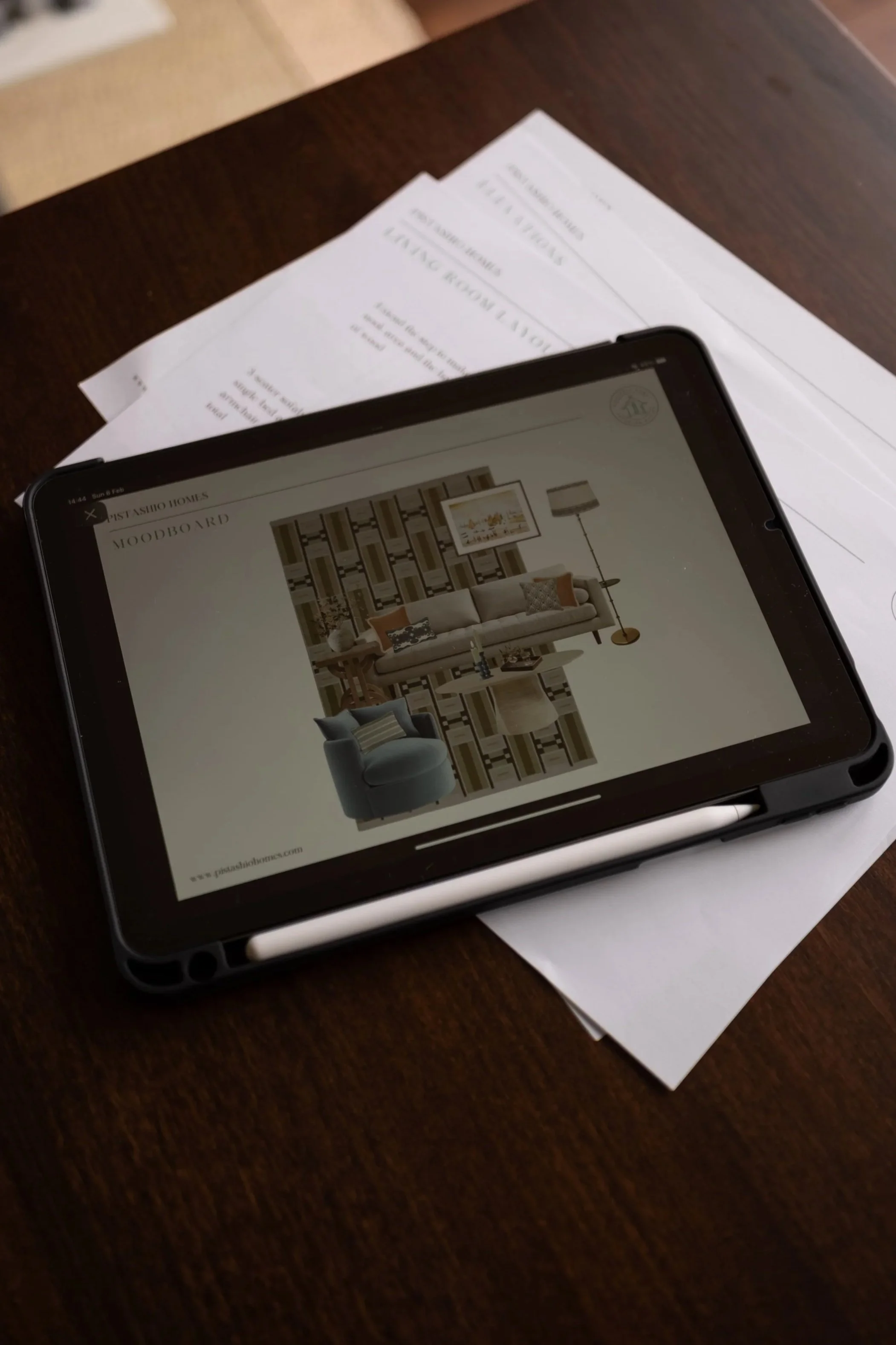 A tablet on a wooden table displaying a furniture catalog, with printed sheets underneath it.
