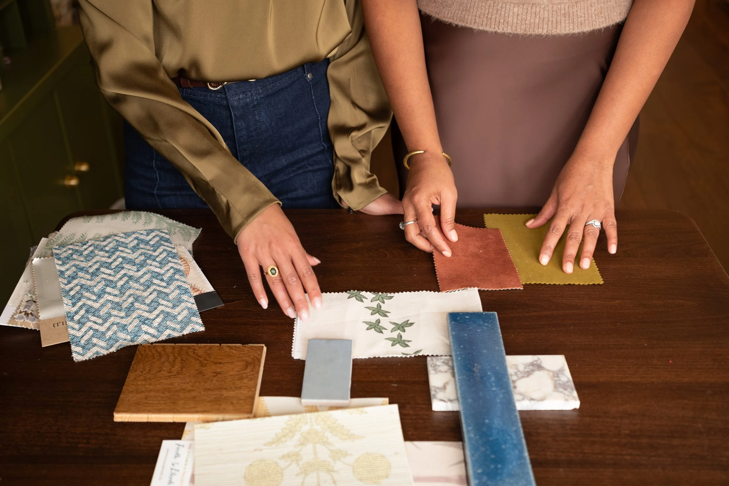 Two women at a table reviewing various fabric and tile samples. One woman wears a tan sweater and mauve skirt with gold jewelry, the other wears a shiny beige blouse and jeans.