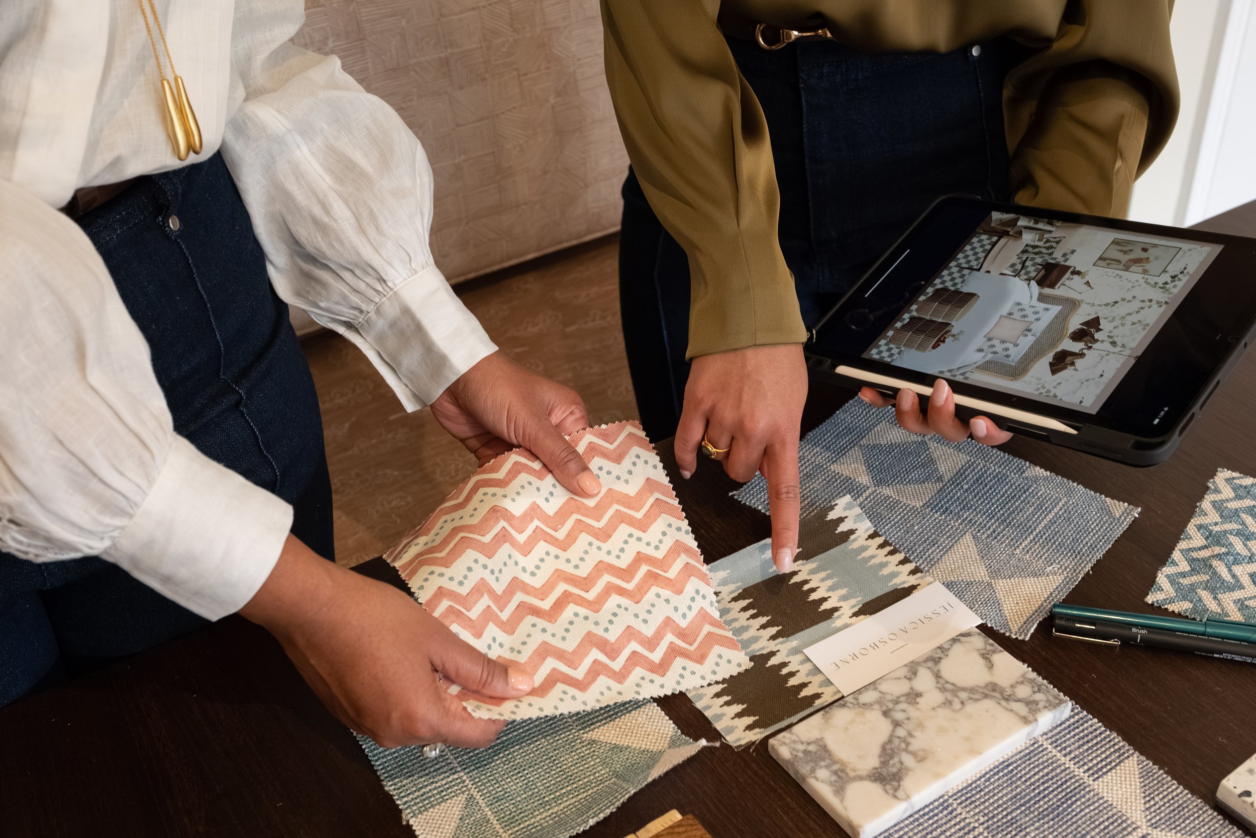 Two people are examining fabric samples and a tablet displaying interior design images on a dark wooden table. One person is pointing at a fabric sample with peach and white zigzag patterns.