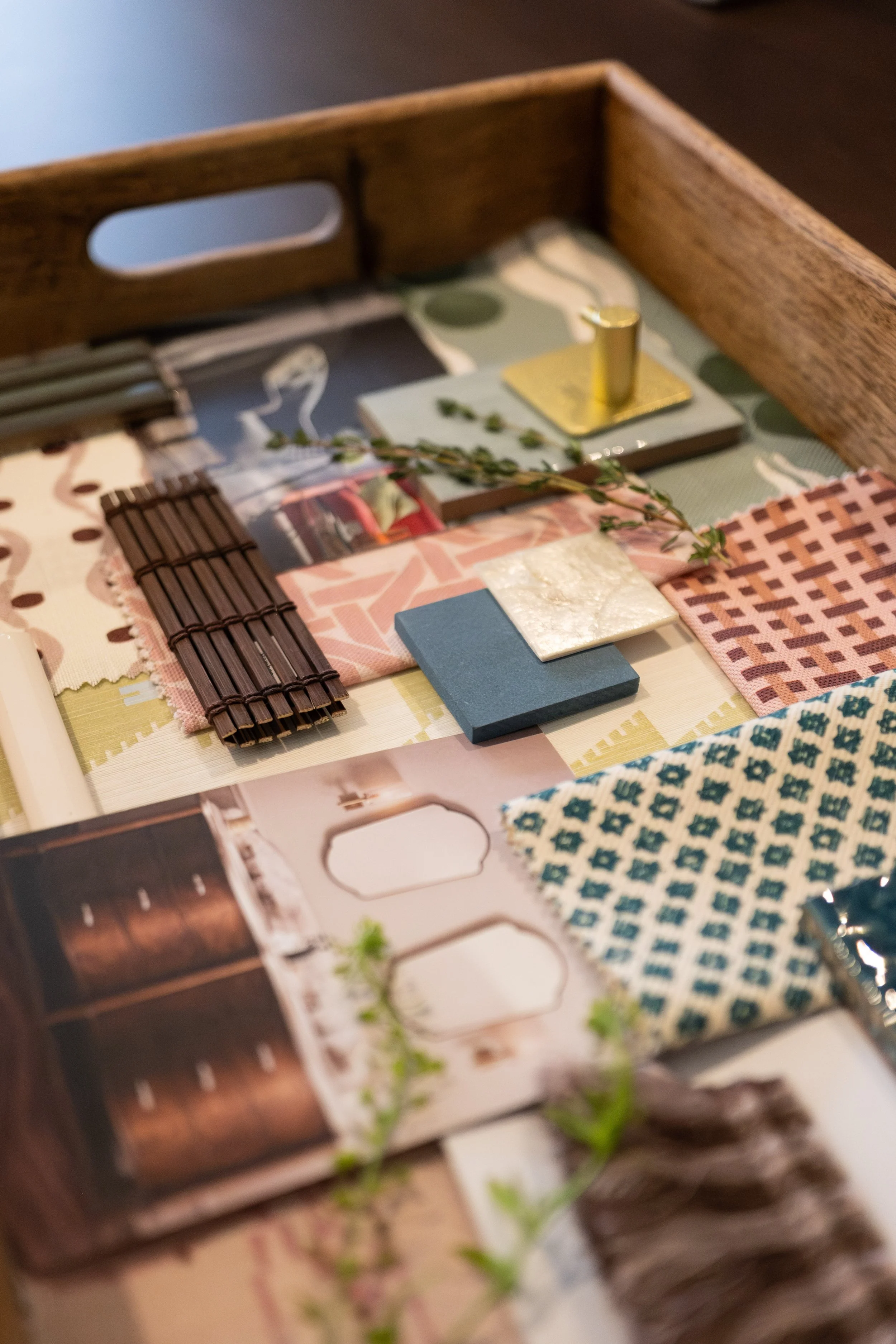 A wooden tray with various patterned paper fabrics, small tile samples, a green sprig, and a metallic pin on a green and white patterned background.