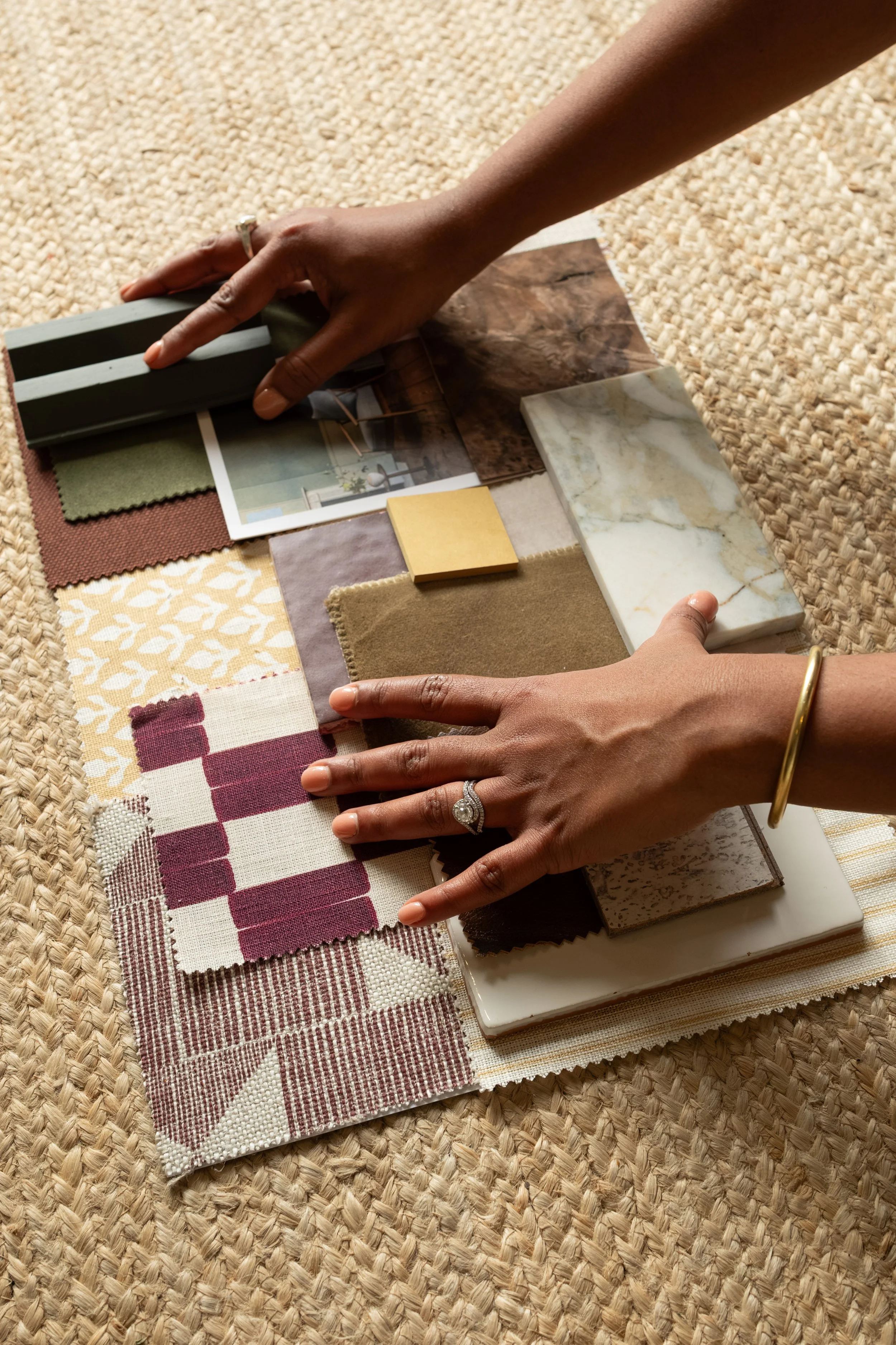 Close-up of hands examining various fabric samples, tiles, and color swatches, laid out on a woven surface.