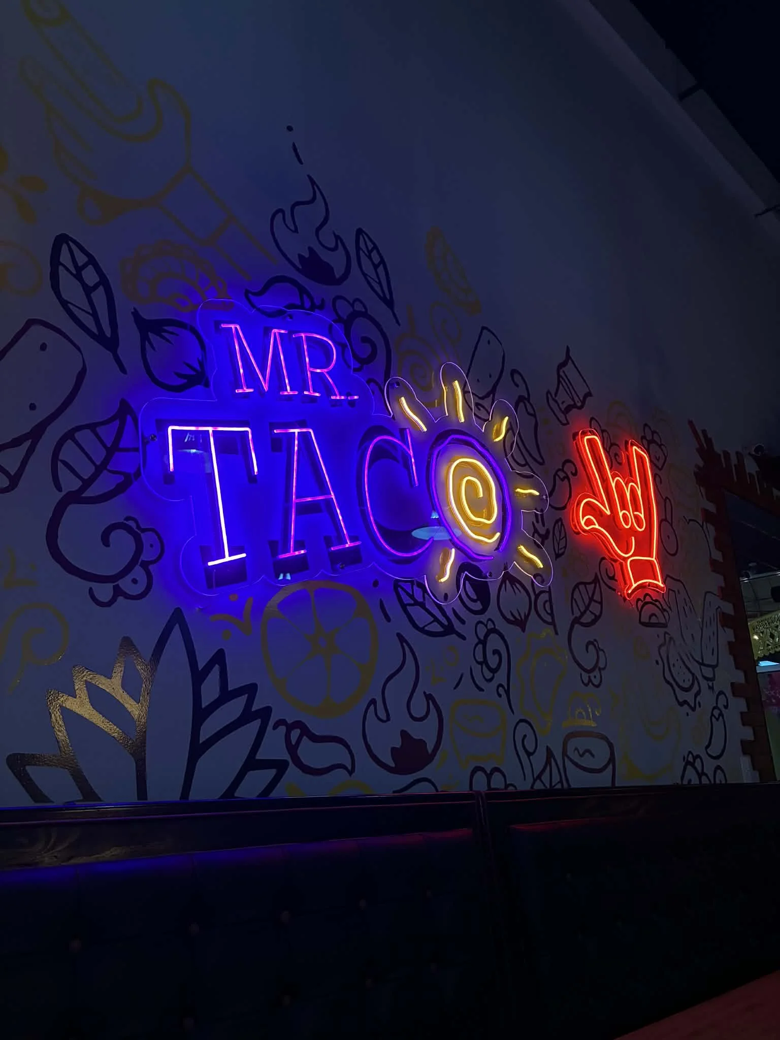 Neon sign that reads 'Mr. Taco' with decorative drawings of leaves and flames on a wall.